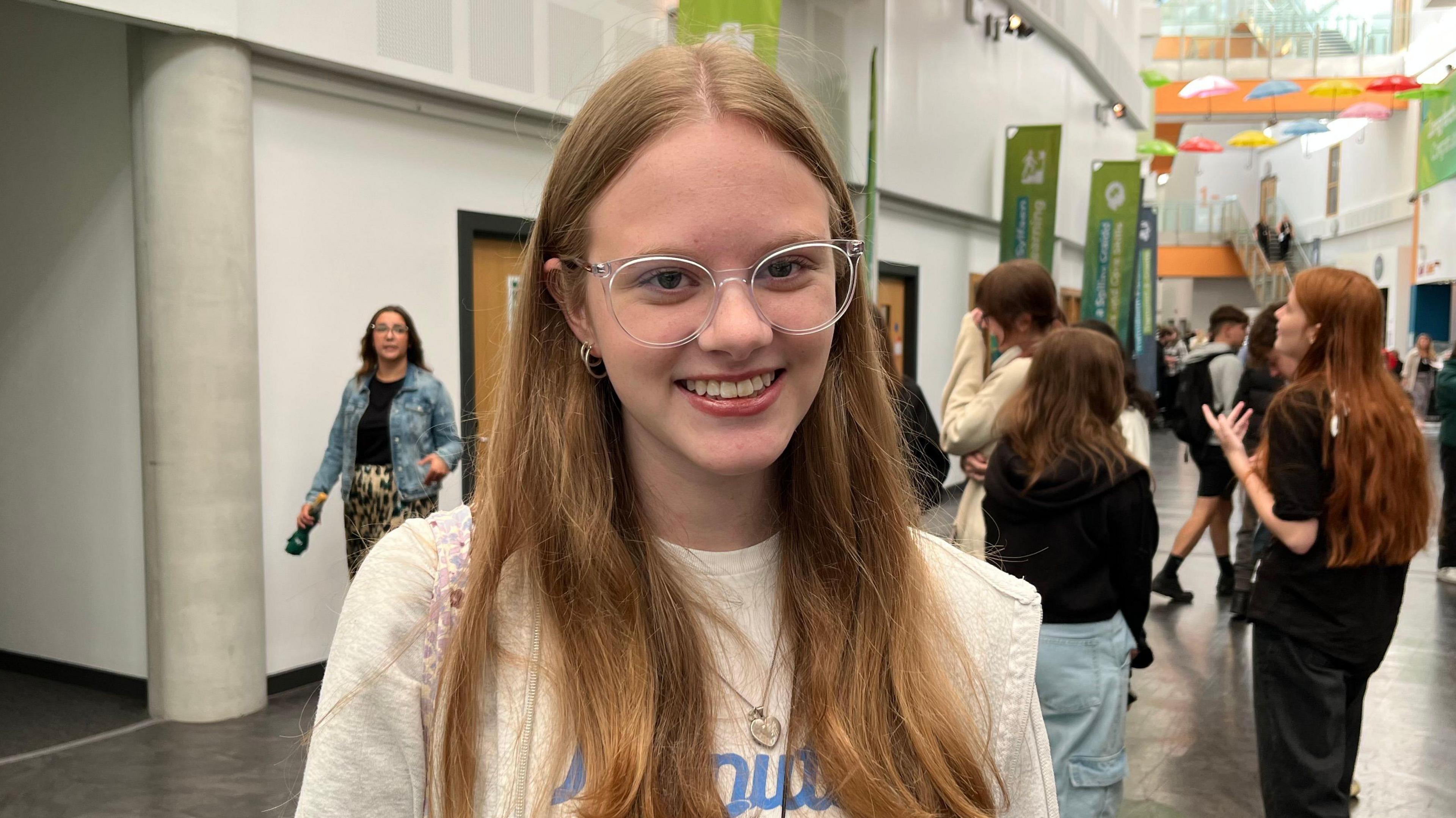 Connie Dolloway, 17, pictured in the college corridor. She wears clear square framed glasses and smiles with her teeth at the camera. She wears a white t-shirt and a grey zip up jacket. She has ginger mid length hair.