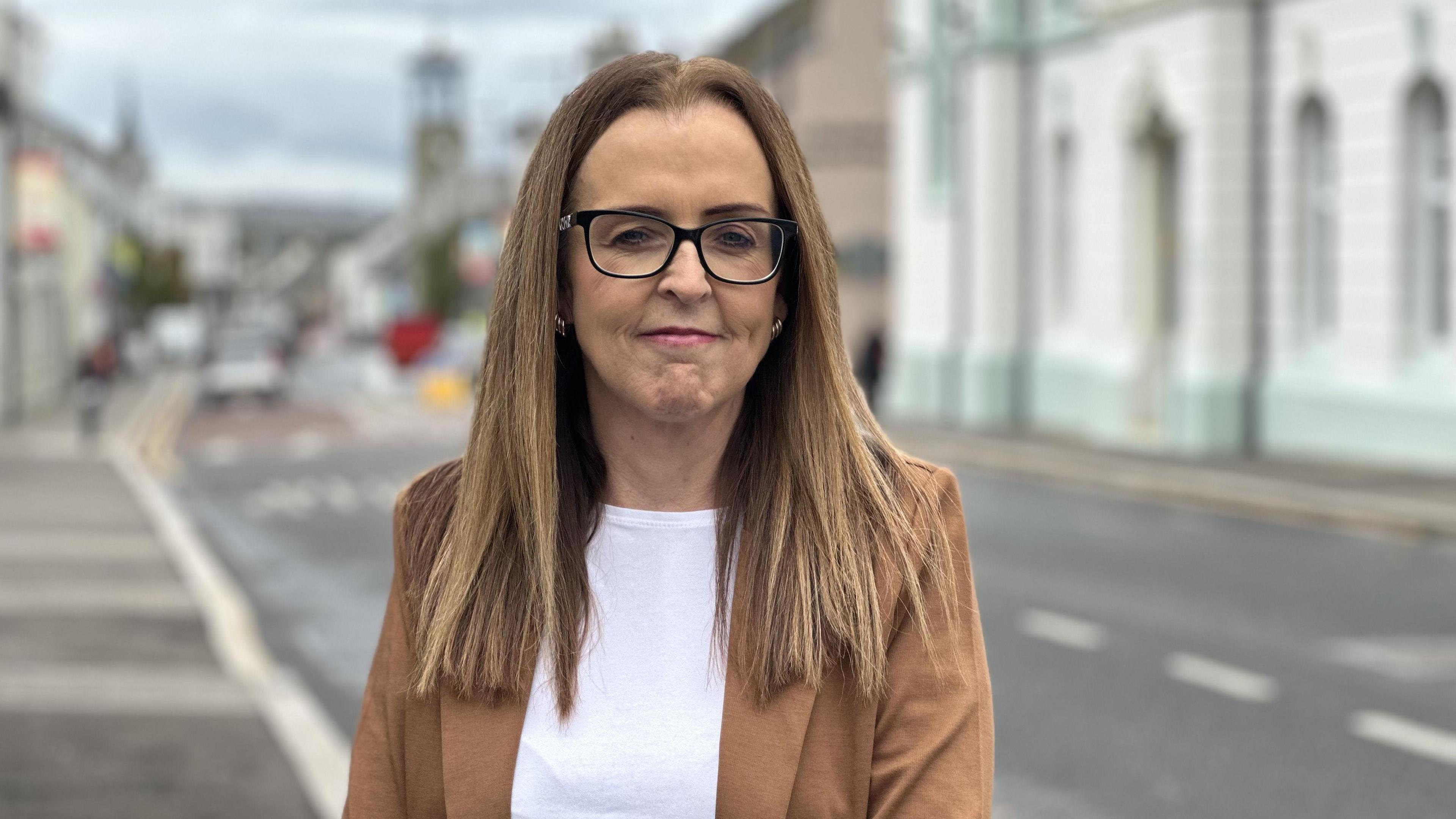 Councillor Cara McShane is standing outside Ballymoney town hall which is out of focus. She is wearing a tan blazer and white tee-shirt. Cara is wearing glasses and has long light brown hair. She is smiling.