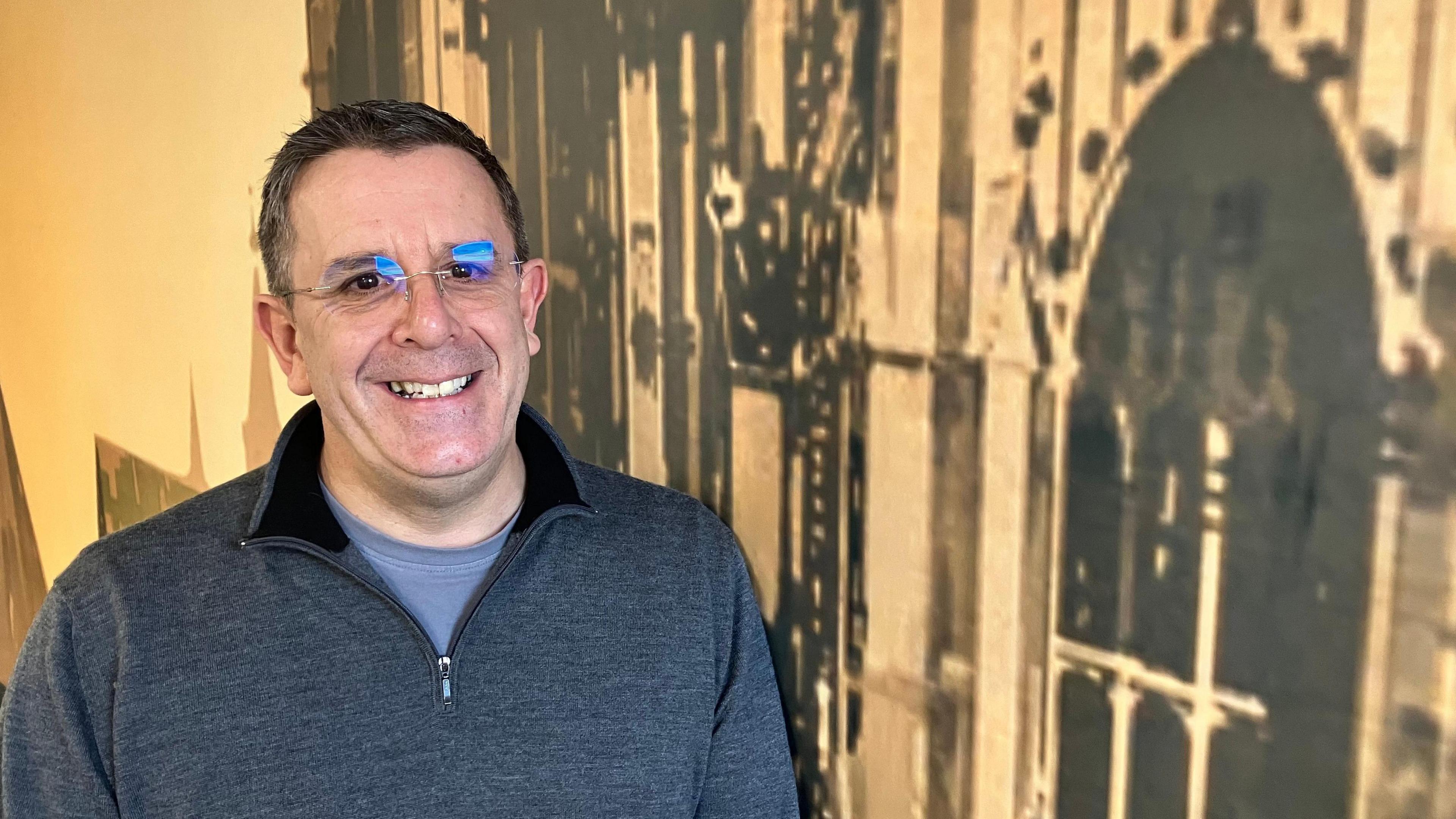 A man with brown hair wears glasses and smiles. He wears a grey jumper.