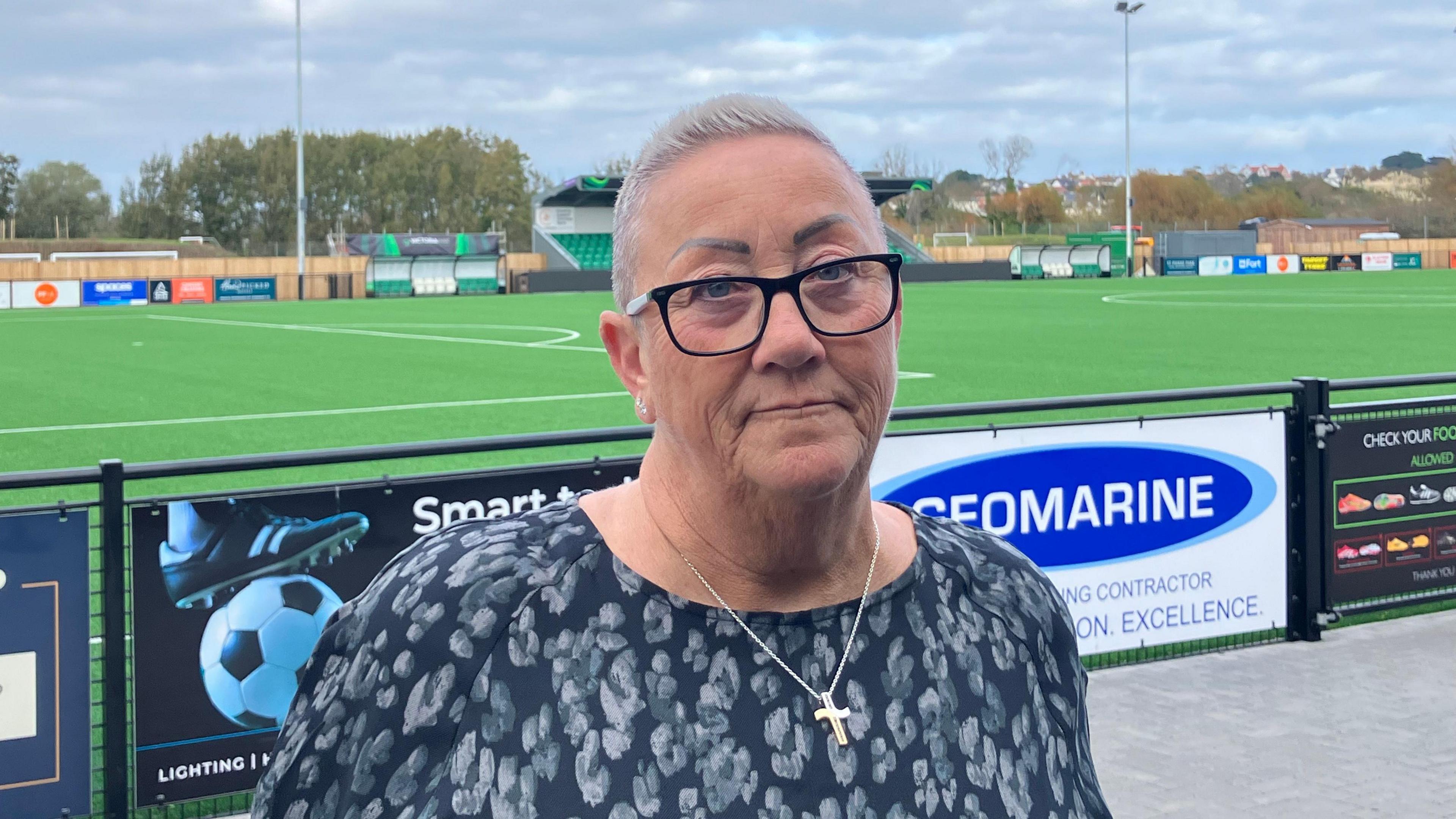 A woman, wearing glasses, looking at the camera. She is waring a blue top and a gold cross necklace. Behind her is a football pitch. The sky is grey and cloudy.