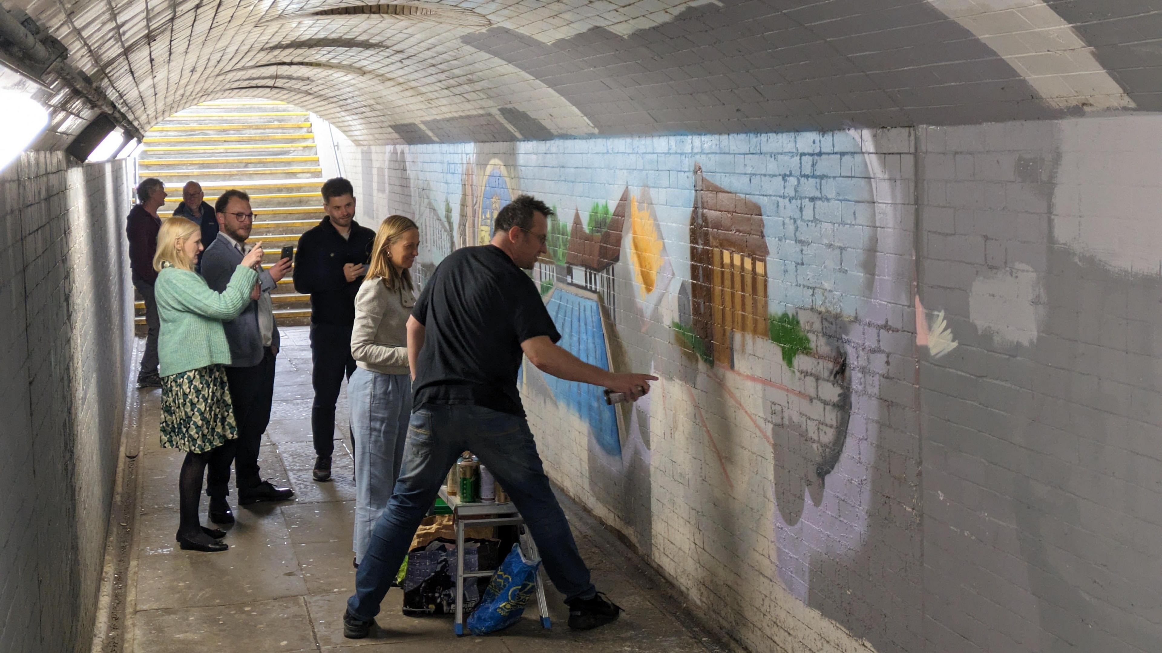 a small crowd of people watch a man spray paint a mural onto a wall in a white and grey tiled underpass