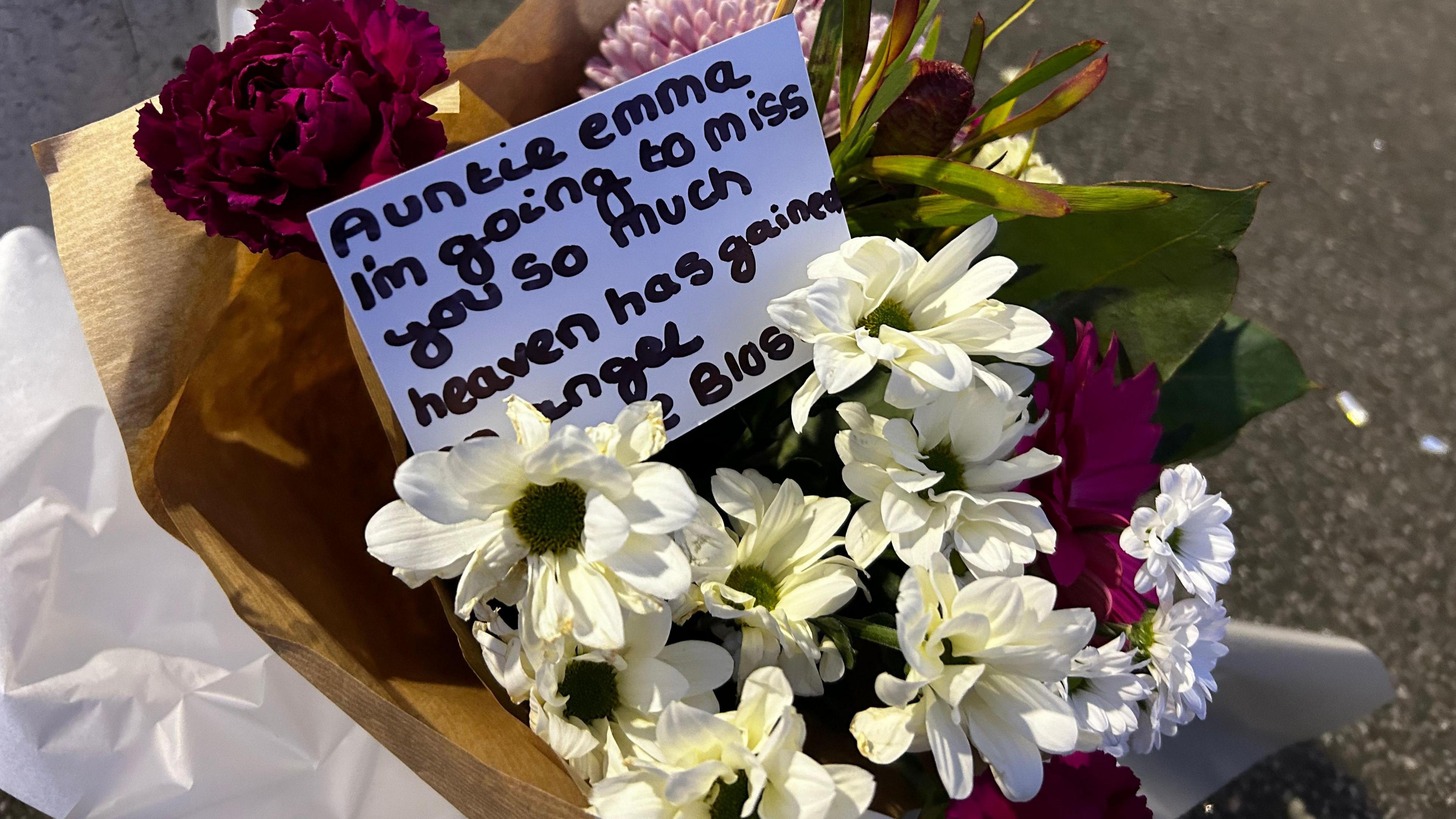 White and pink flowers with a card that reads "Auntie Emma, I'm going to miss you so much. Heaven has gained an angel."