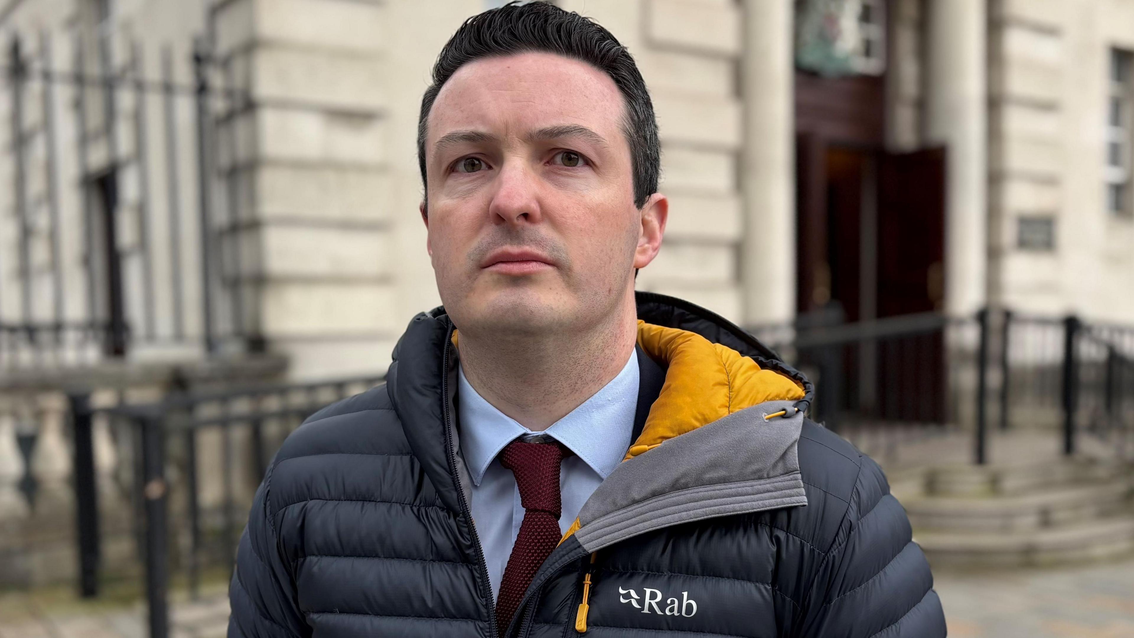 John Patrick Clayton outside court. He is wearing a grey puffer jacket, blue shirt and burgundy tie. He has black hair.