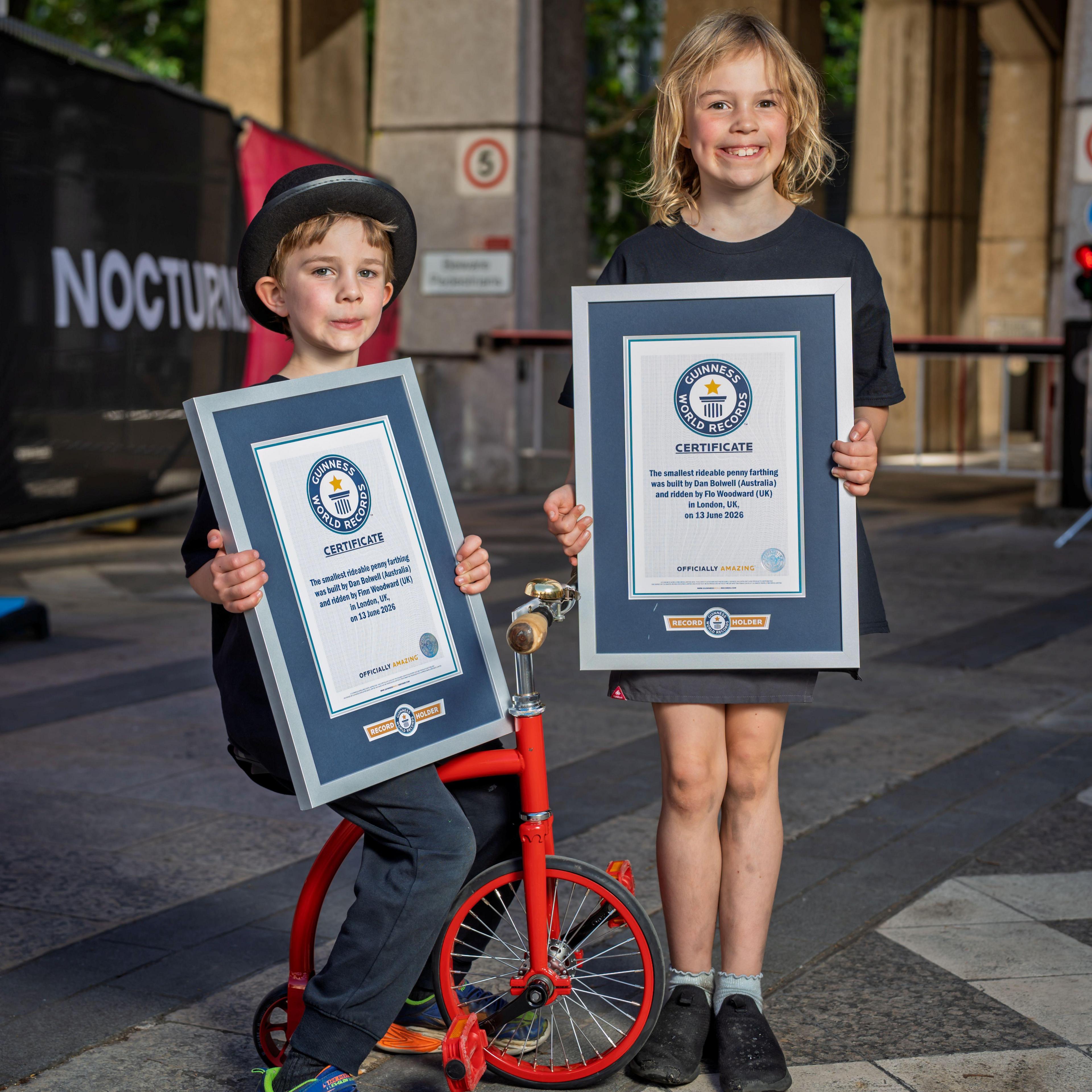 a boy and girl with a small red penny farthing smile into the camera holding framed Guinness world record certificate. the boy is sat on the bike whilst the girl stands. the backdrop is a building, unknown, in London.