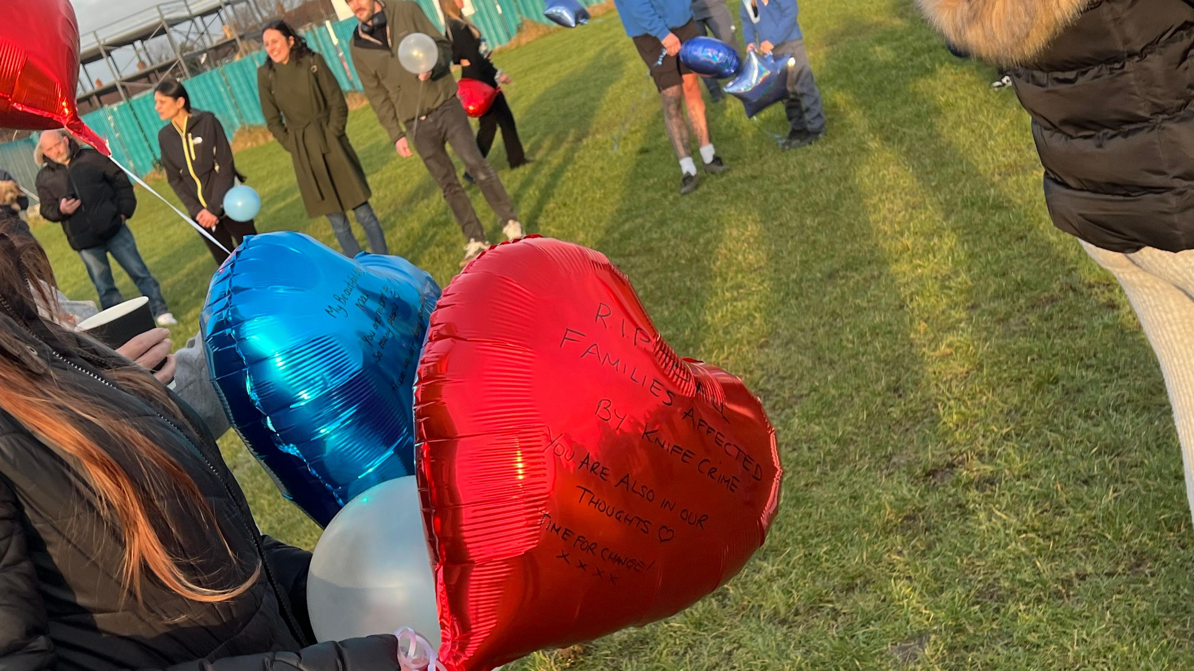 A woman standing on a field at sunset as part of a gathering of people holds a red heart shaped balloon which reads "RIP to all families affected by knife crime. You are also in our thoughts. Time for change". 