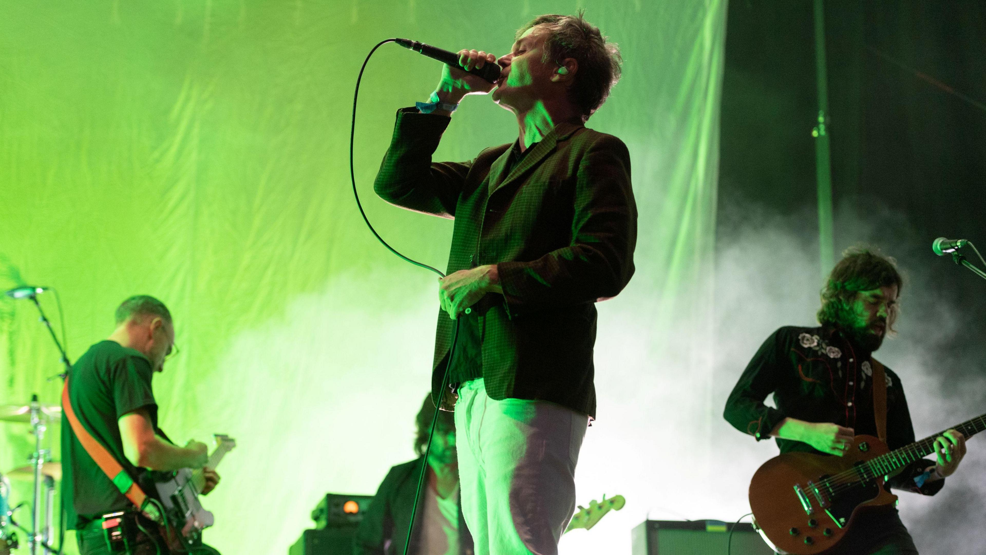 A band performing onstage - the singer has the microphone raised to his mouth, while two guitarists and the bassist are visible in the background. Green/yellow lighting covers the stage.