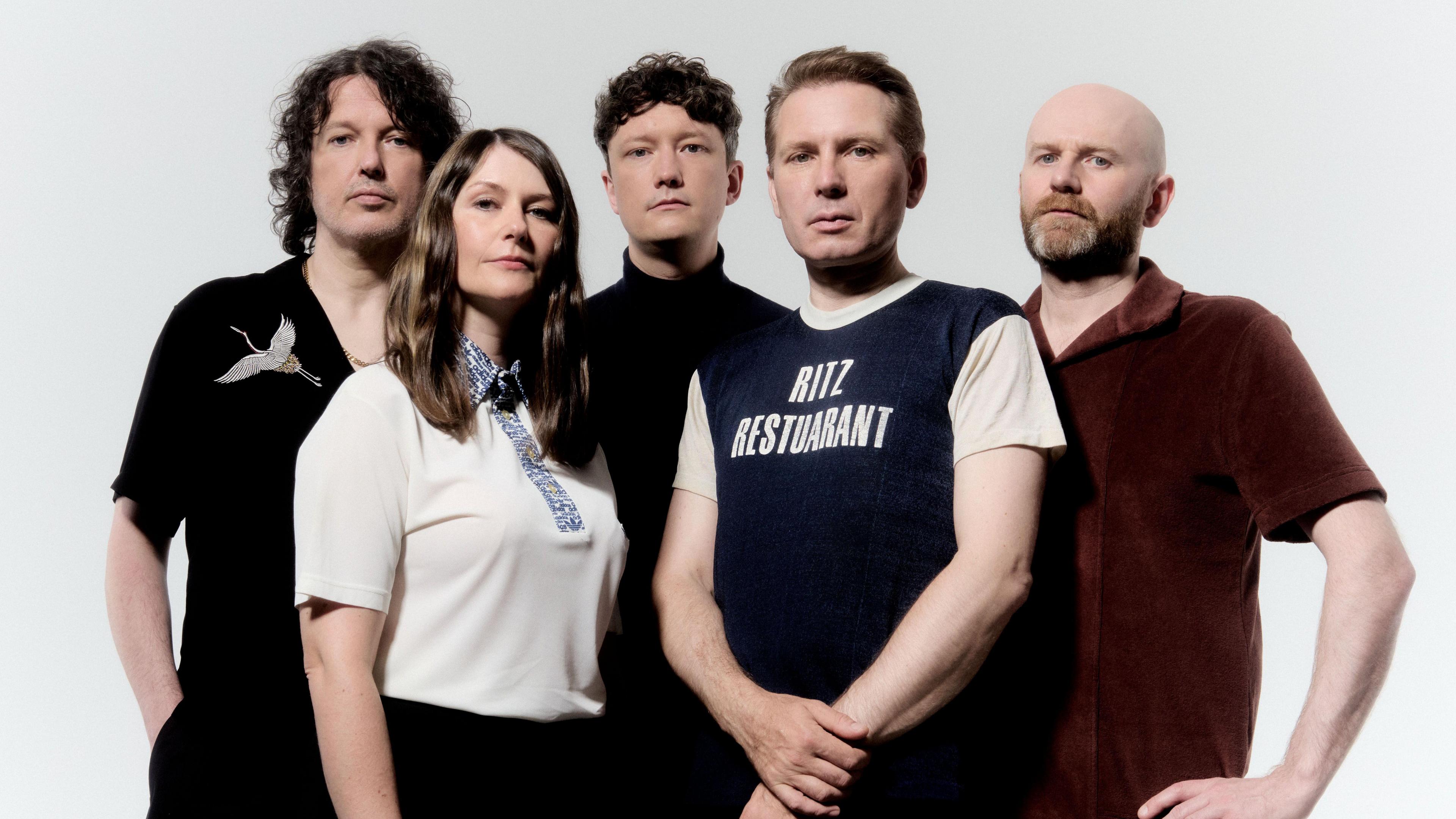 Franz Ferdinand look into the camera. Alex Kapranos is wearing a top with RITZ RESTUARANT written on it. The group are dressed casually, in shirts of varying colours (black, red and white). 