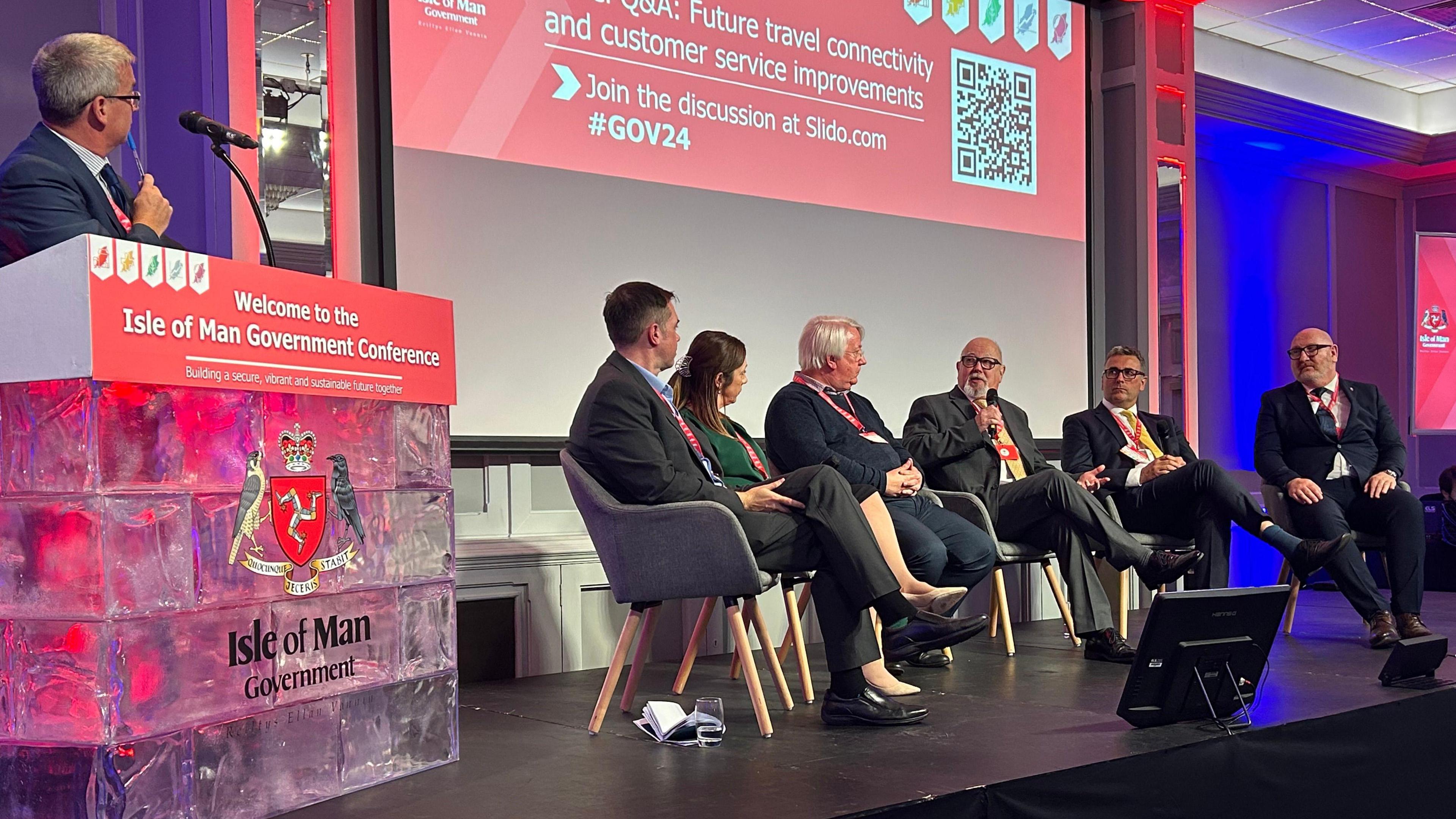 Five men and a women sit in a semi circle on chairs on a stage. One is speaking into a microphone. There are large red signs in the background with the Government Conference branding and another man is standing by a lectern holding a pen to his mouth.