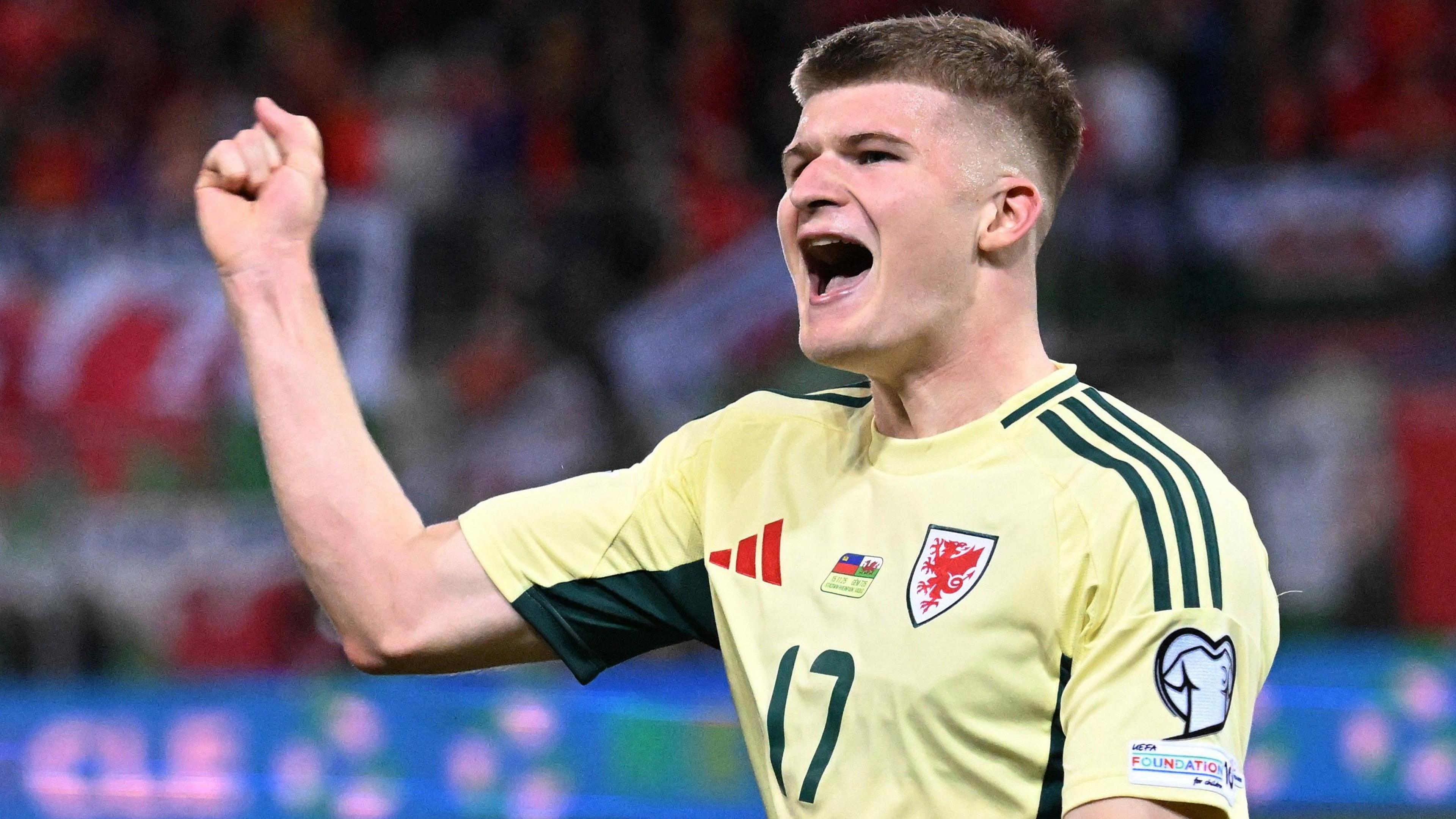 Jordan James punches the air and celebrates after scoring for Wales against Liechtenstein. He is wearing a light yellow shirt with dark green trim.