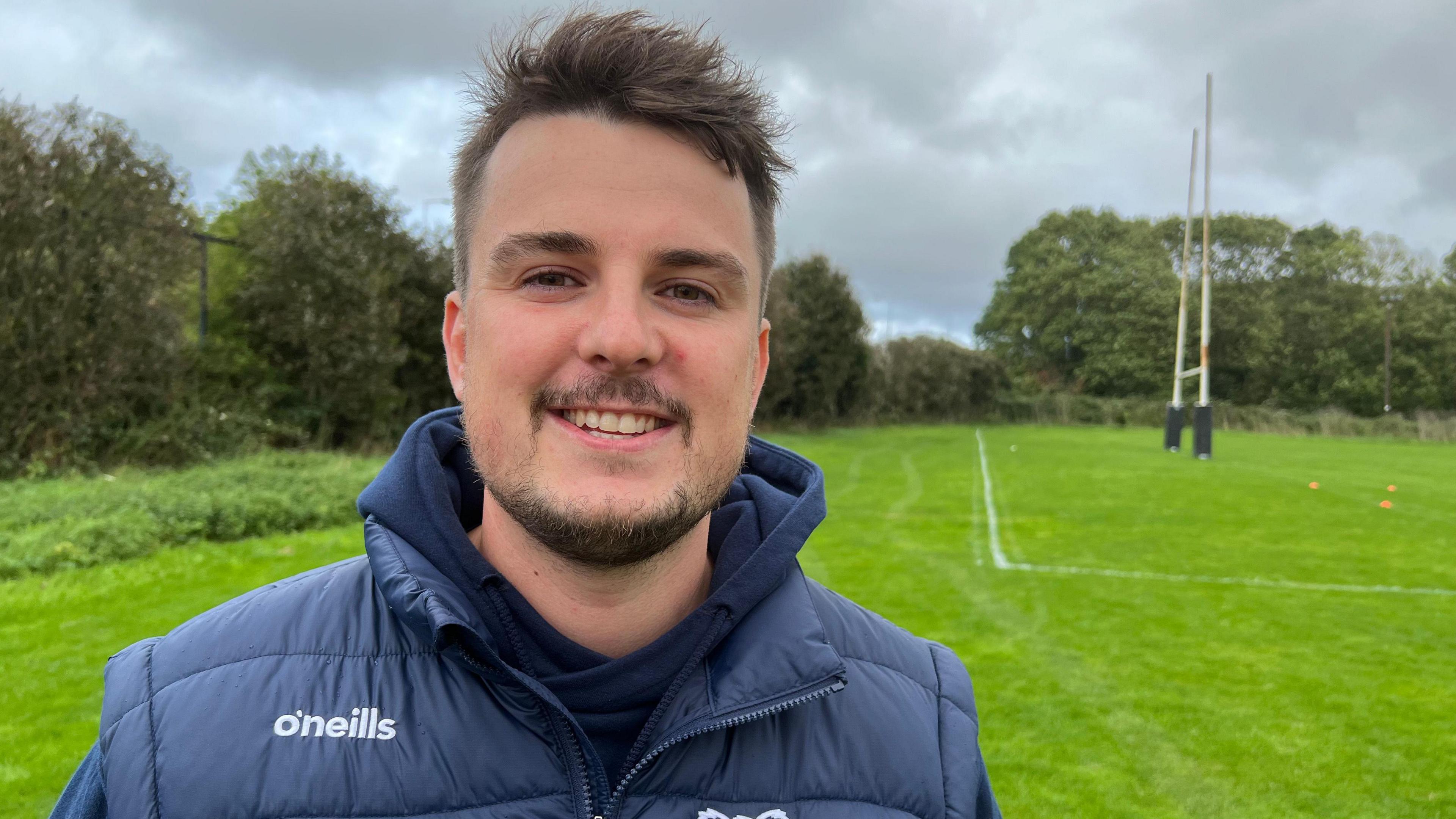 Jordan Lovatt wearing a padded navy body warmer and hoodie. He is standing on a rugby pitch with the goal visible in the background, smiling at the camera. He has a short goatee style beard and brown hair, which is longer on the top.