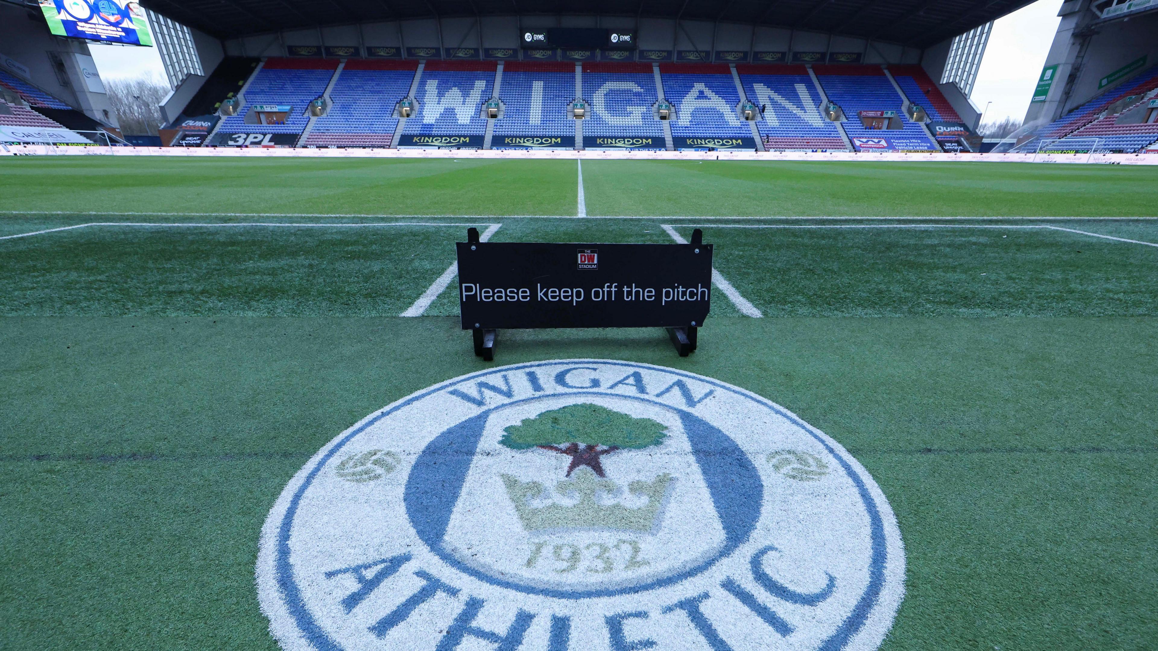 A view of Wigan's pitch with a Wigan Athletic badge on the artificial turf in front of the pitch and 'WIGAN' on the seats in the stand on the far side of the pitch    