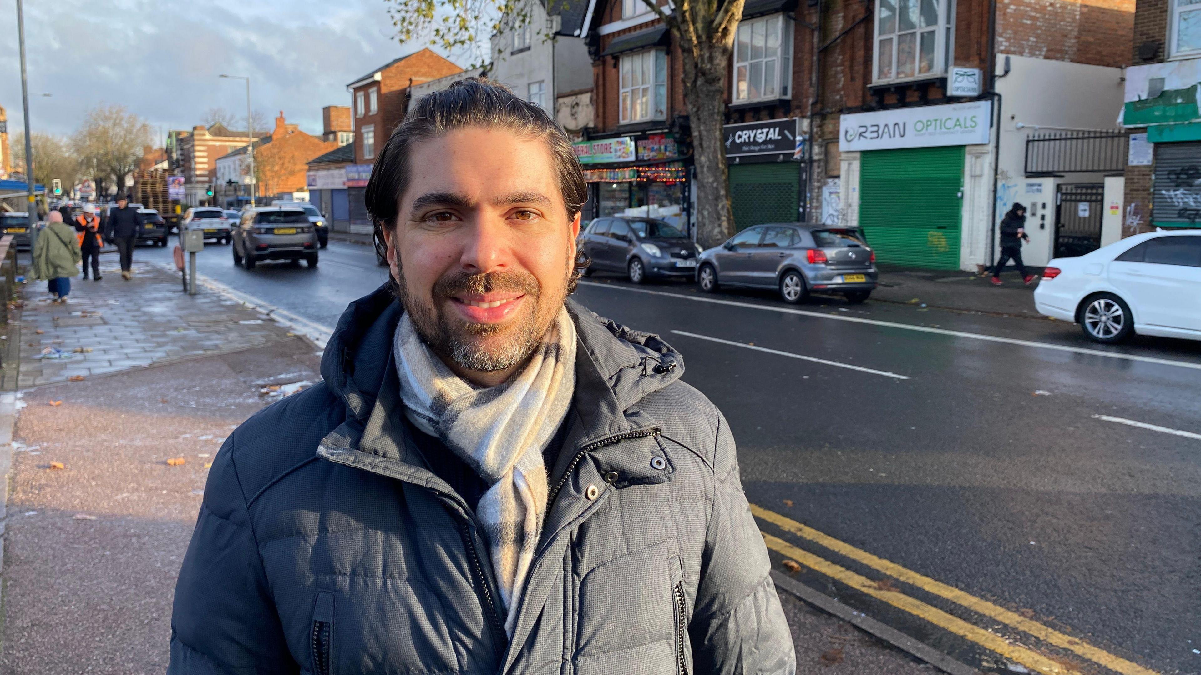 Dr Danilo Spinola pictured with slicked-back black hair. He is smiling and wearing a grey hooded coat and tartan print scarf.