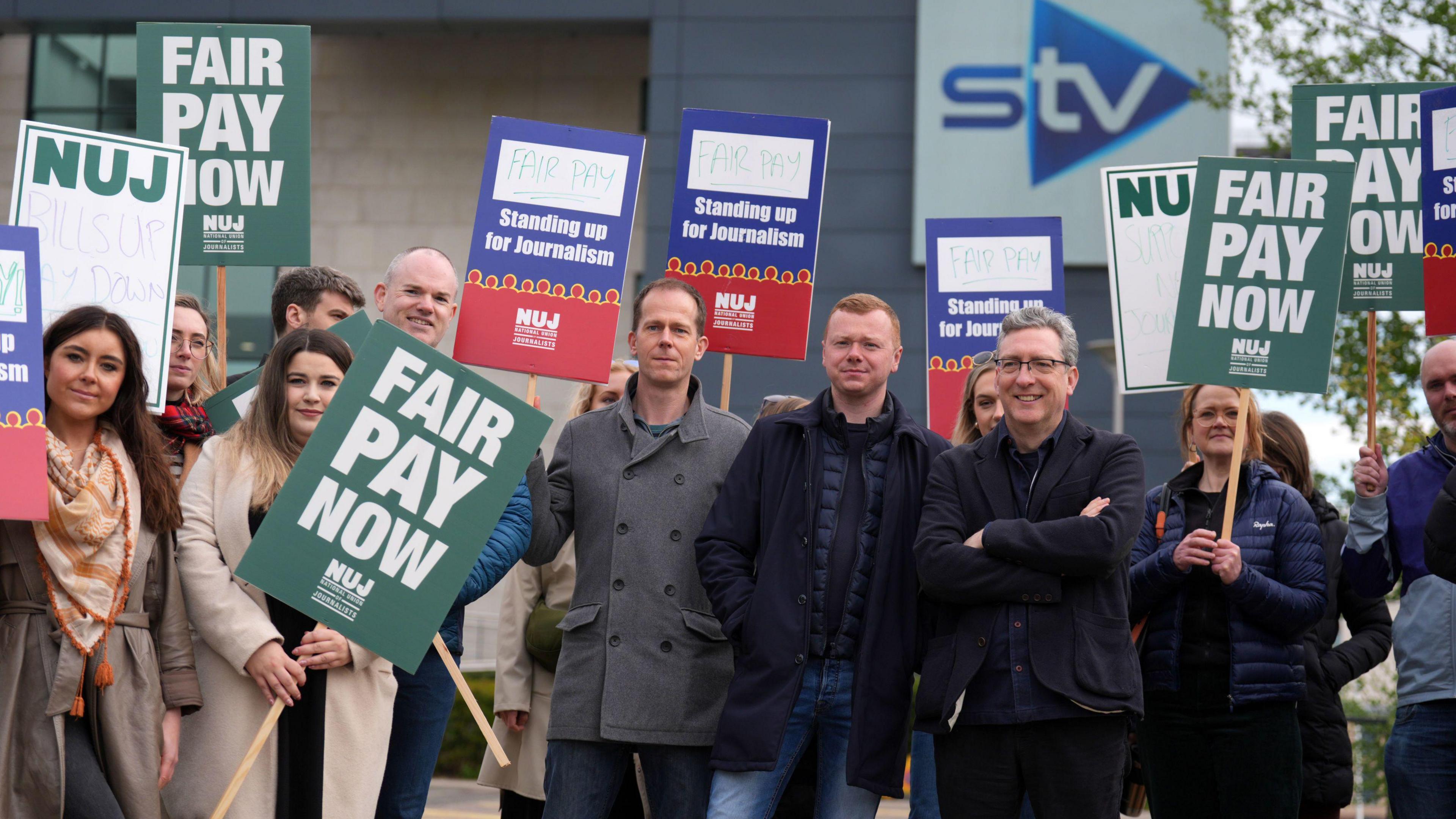 A group of people holding placards outside a building with the STV logo. Signs read “Fair Pay Now” and “Standing up for Journalism,” representing a protest organized by the National Union of Journalists (NUJ).