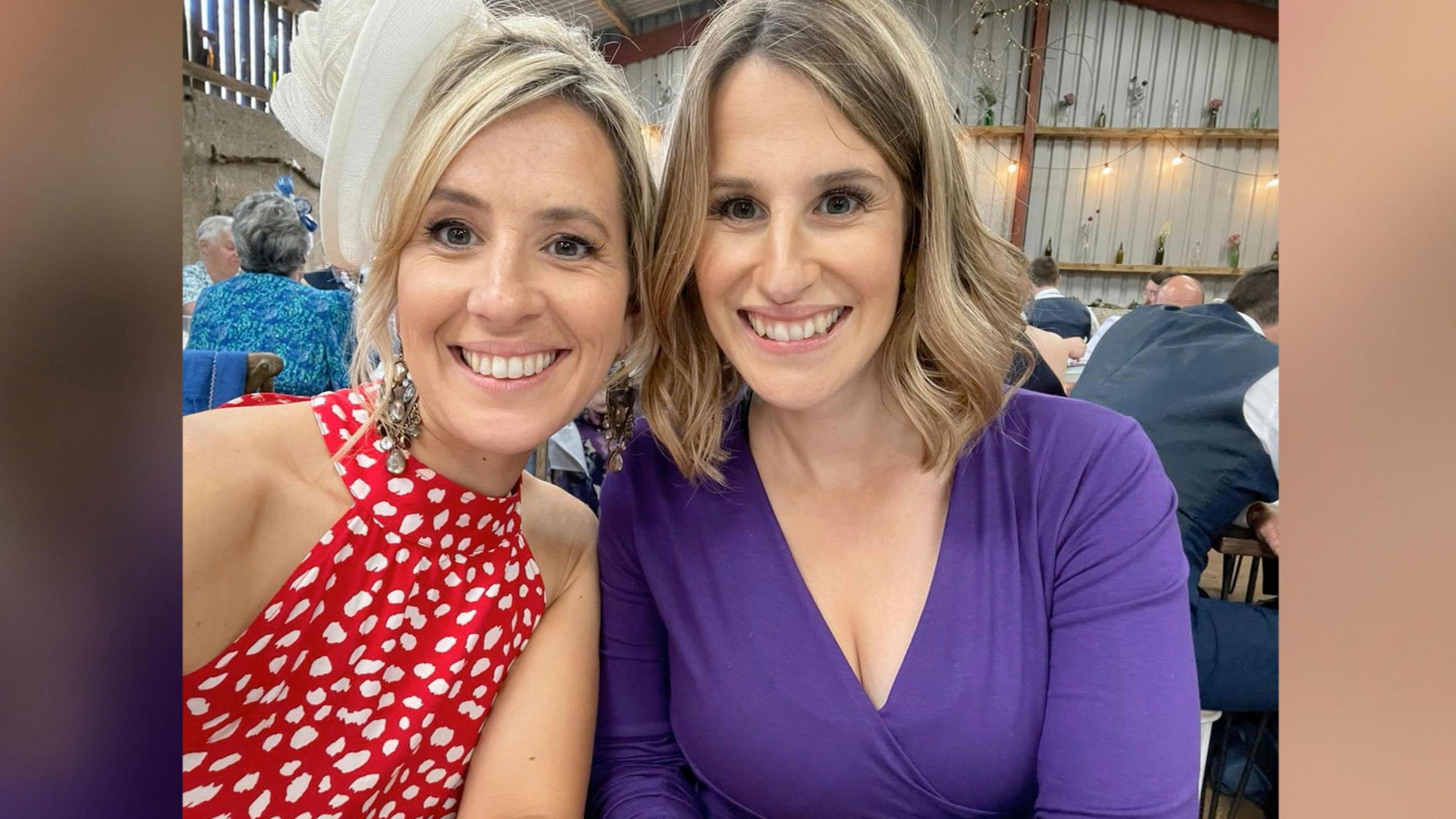 Mari (left) is wearing a red and white polka dot halter neck dress with a white fascinator in her blonde coloured hair, which is tied back. Her sister, Lisa, (right), is wearing a long sleeve purple dress and her short blonde hair is curled. They are both smiling in a selfie at a wedding in a barn.