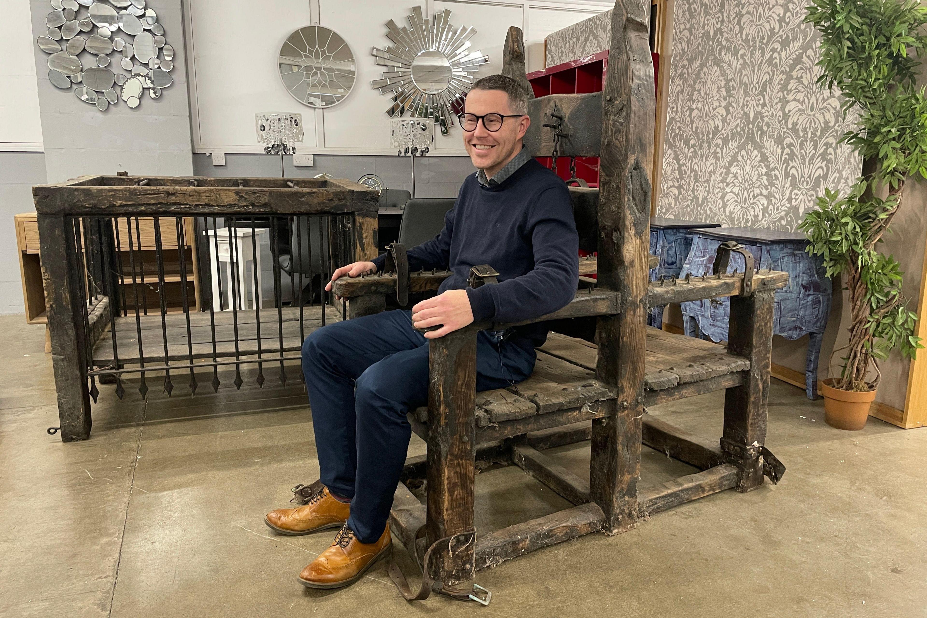 A man sitting in an interrogation chair with a wooden and metal cage in the background
