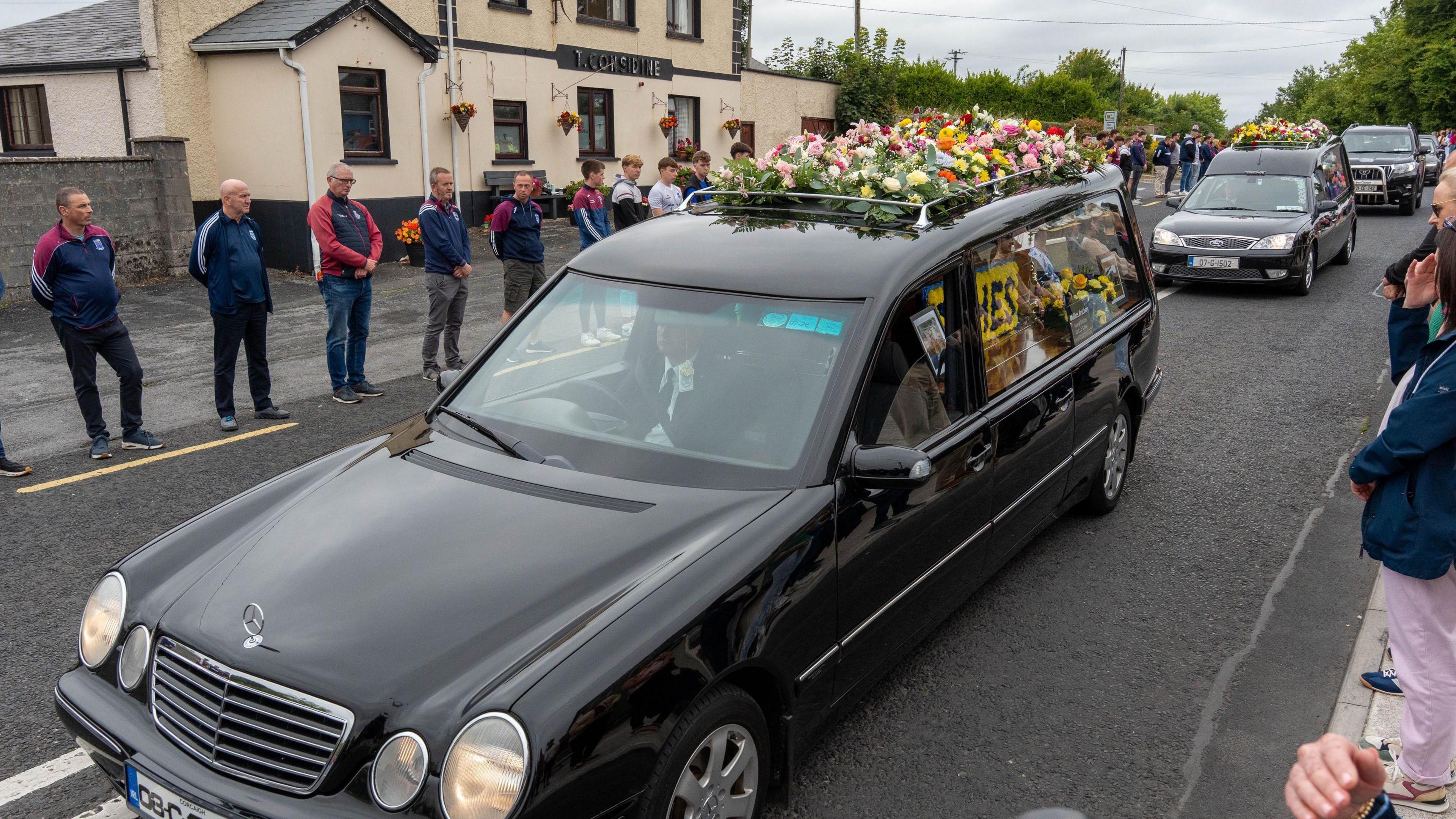 Two black hearses with flowers on top are driving down the street of a small town. A number of people in GAA tops can be seen on the other side of the street, forming a guard of honour.