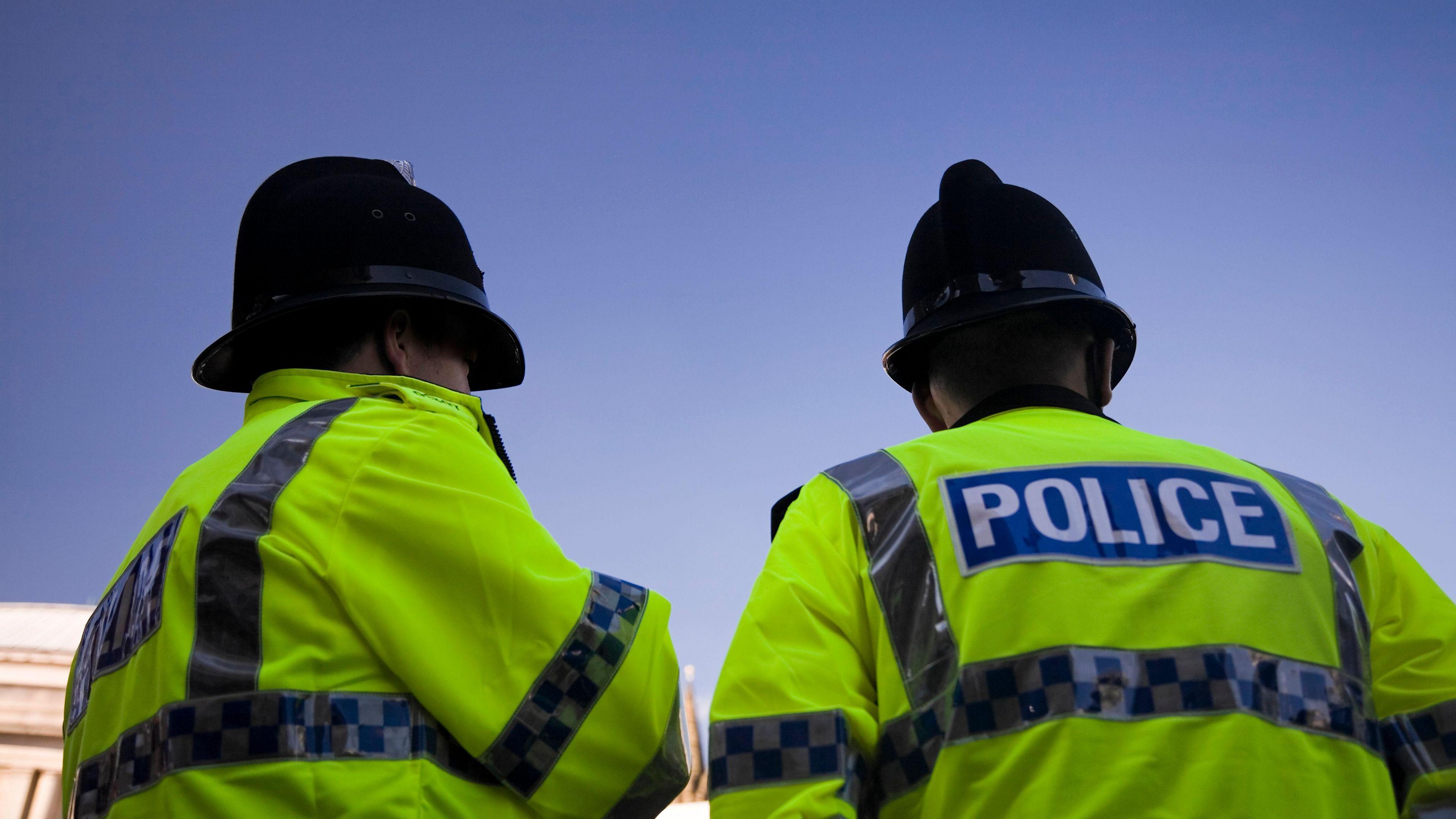 Two police officers in yellow hi vis jackets and with black hats on stand outside a crime scene