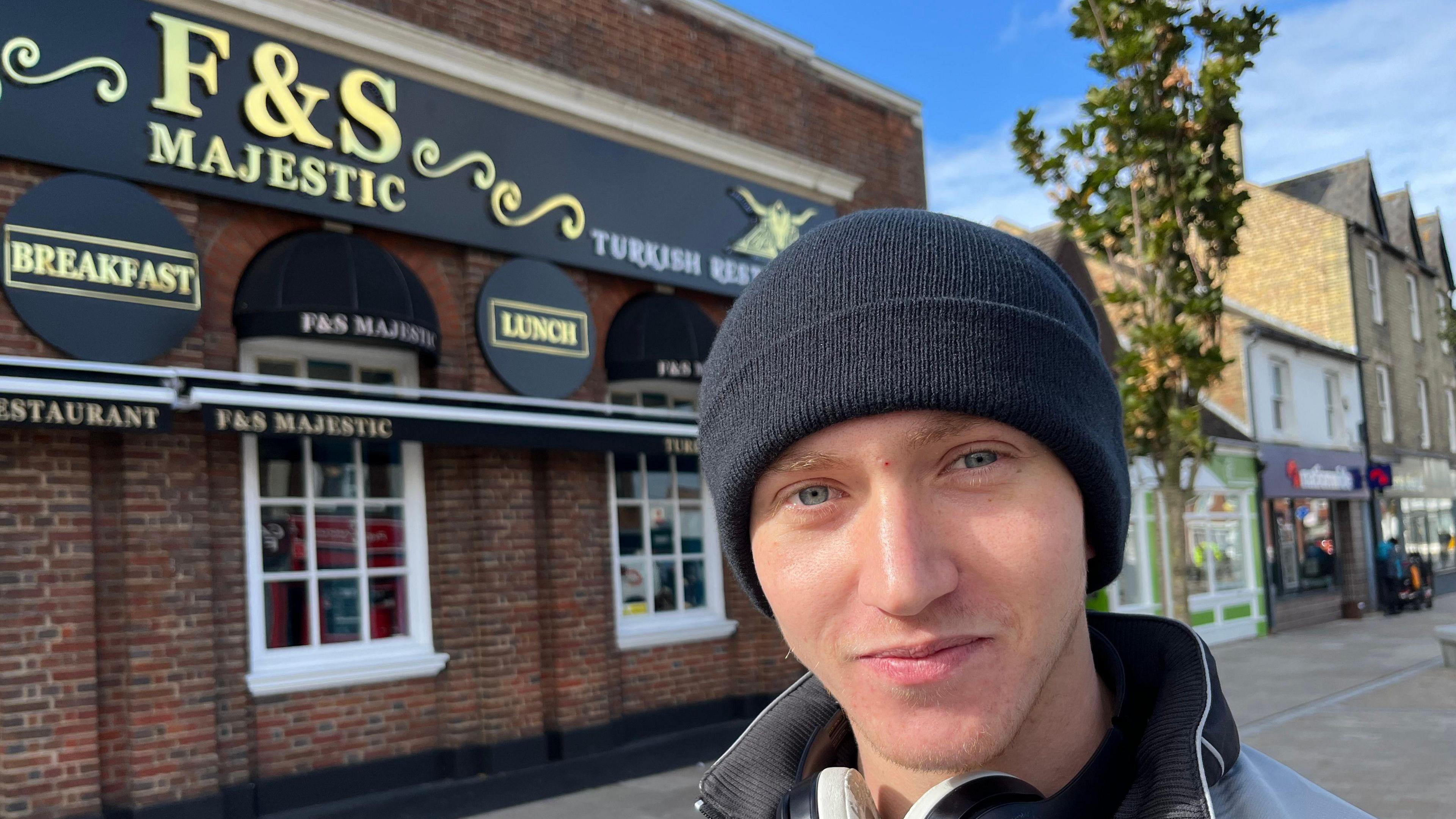 Harry Pluckrose, 19, has a black beany hat on and a black and grey tracksuit top with the collar up, he has wireless headphones around his neck and behind him is the F&S Turkish restaurant sign and building.