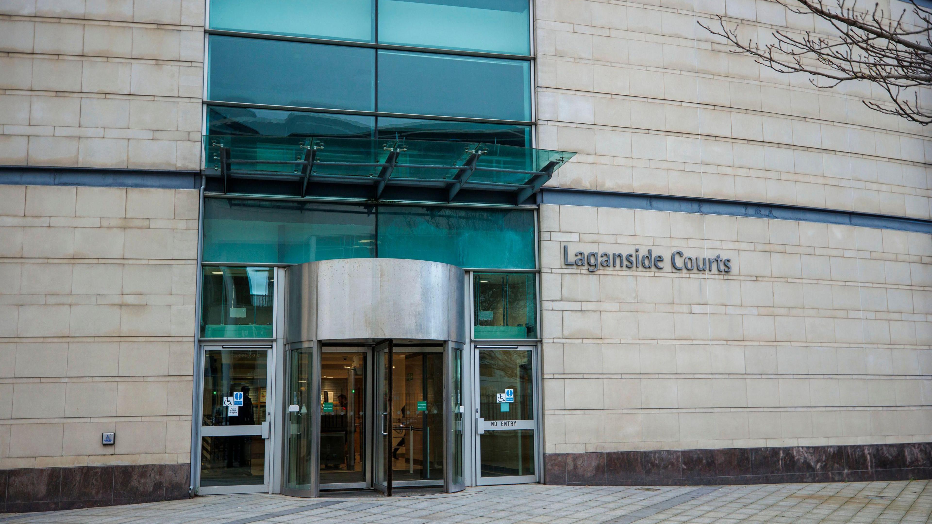 The exterior of Laganside Courts in Belfast. It is a white stone building with a glass panelled entry with a rotating door. To the right of the door is a sign saying 'Laganside Courts' in silver metal.