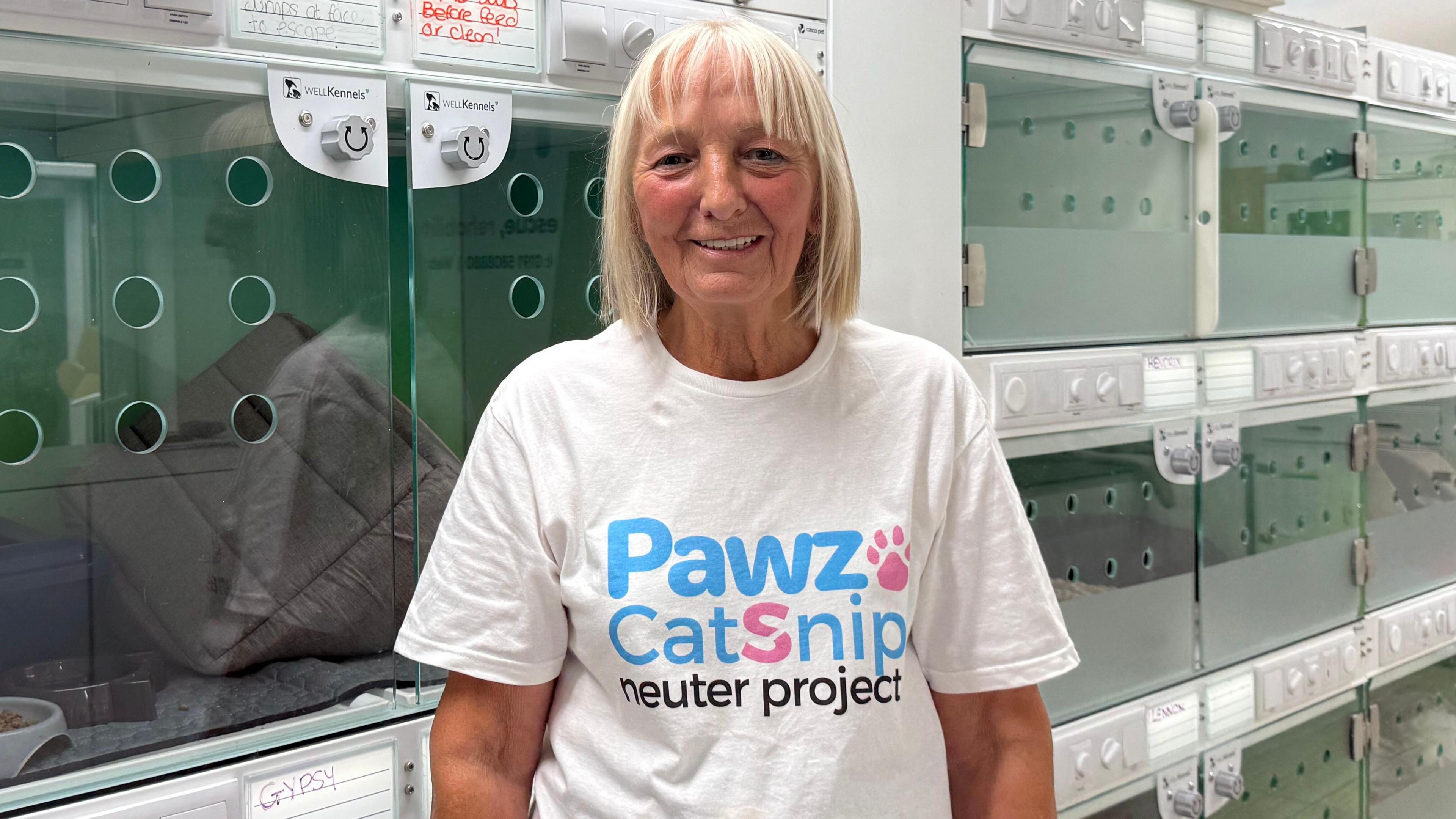 Lynne Ebdale has a blonde bob haircut and is wearing a white T-shirt that reads 'PAWZ CATSNIP NEUTER PROJECT'. She stands in a veterinary practice front of animal chambers where animals have been rescued and treated.