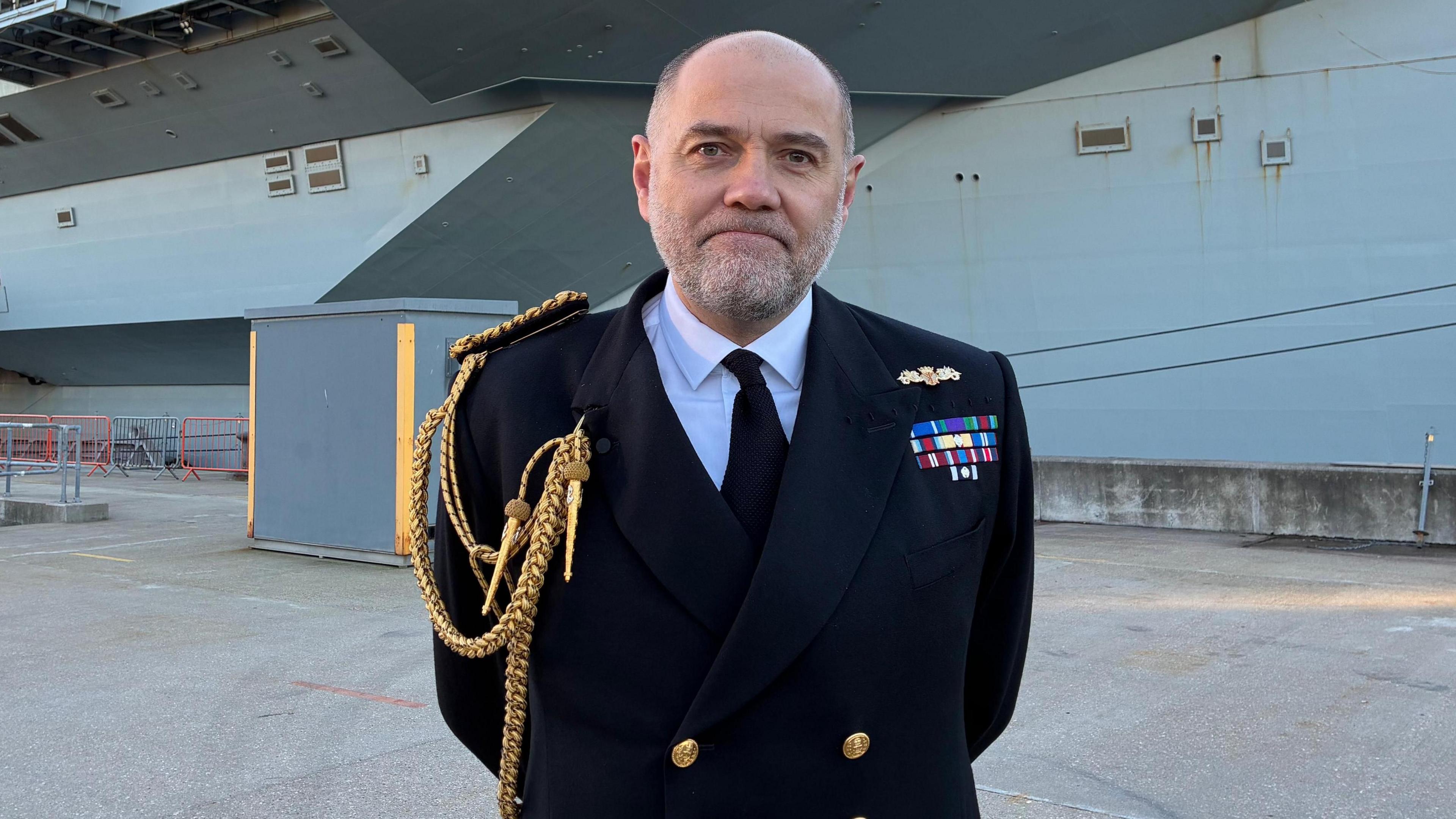 Vice Adm Stephen Moorhouse, CBE stands on Portsmouth Naval Base in front of HMS Prince of Wales. He is wearing a Royal Navy uniform consisting of a darkj blue blazer, with a golden cord trailing down from his left shoulder into his breast pocket. He is bald with short grey stubble.
