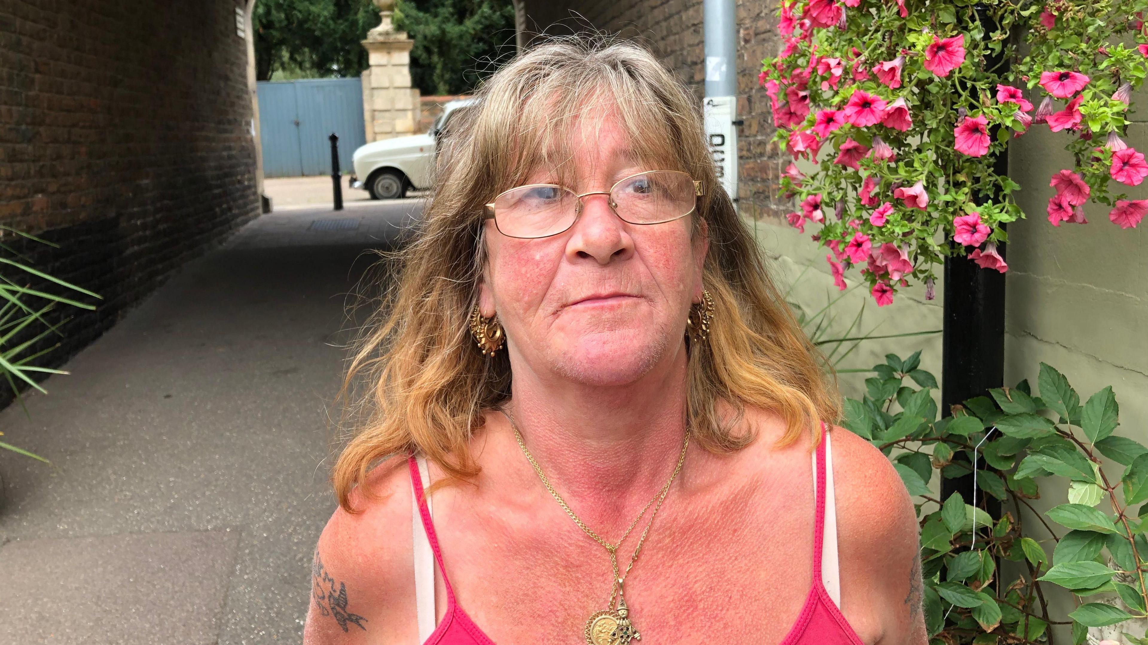 Diane Rowlett wearing a pink and white vest top stands in an alleyway next to pink flowers in a hanging basket.