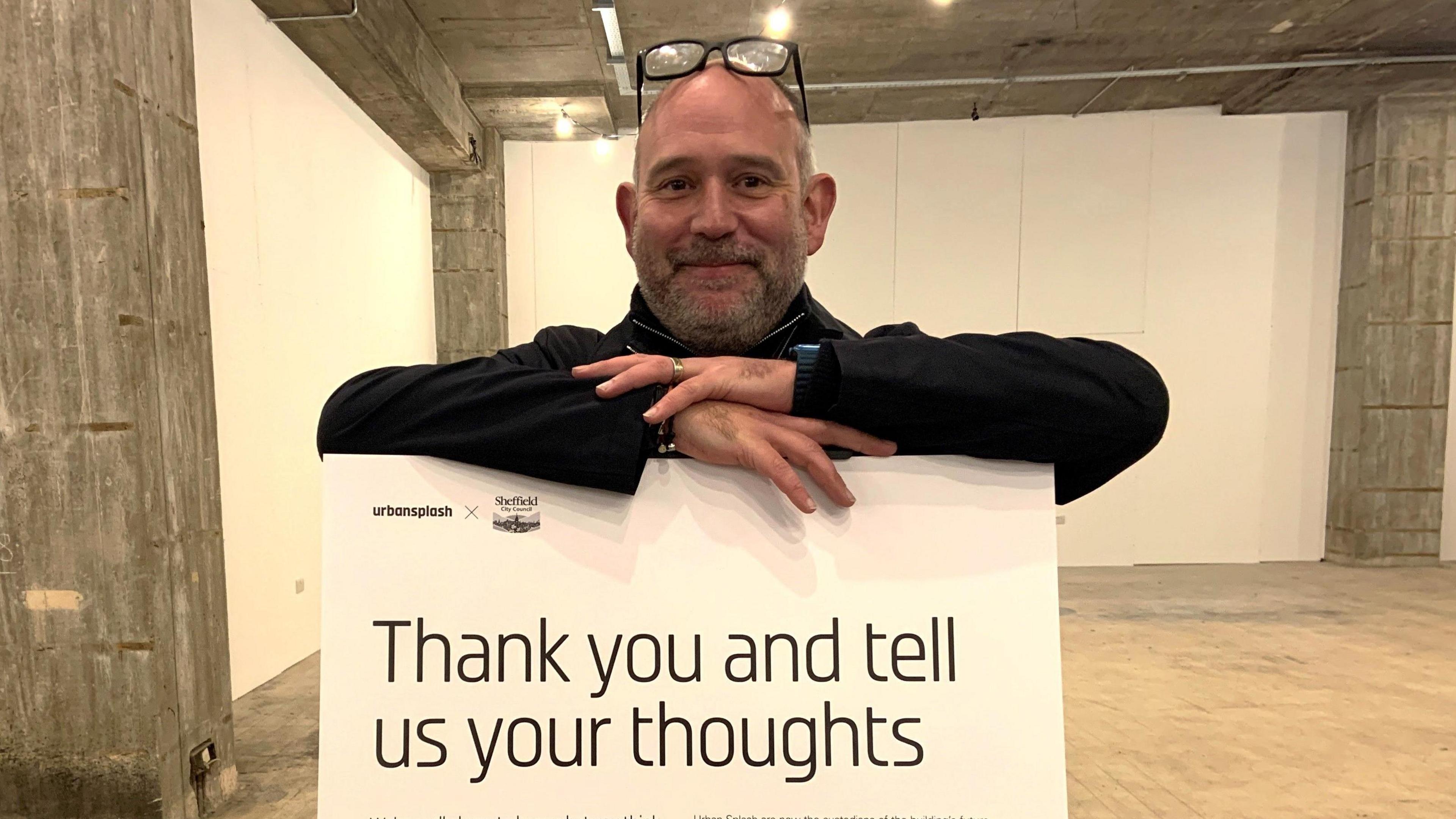 A man in a dark top stands behind a sign saying "thank you and tell us your thoughts"