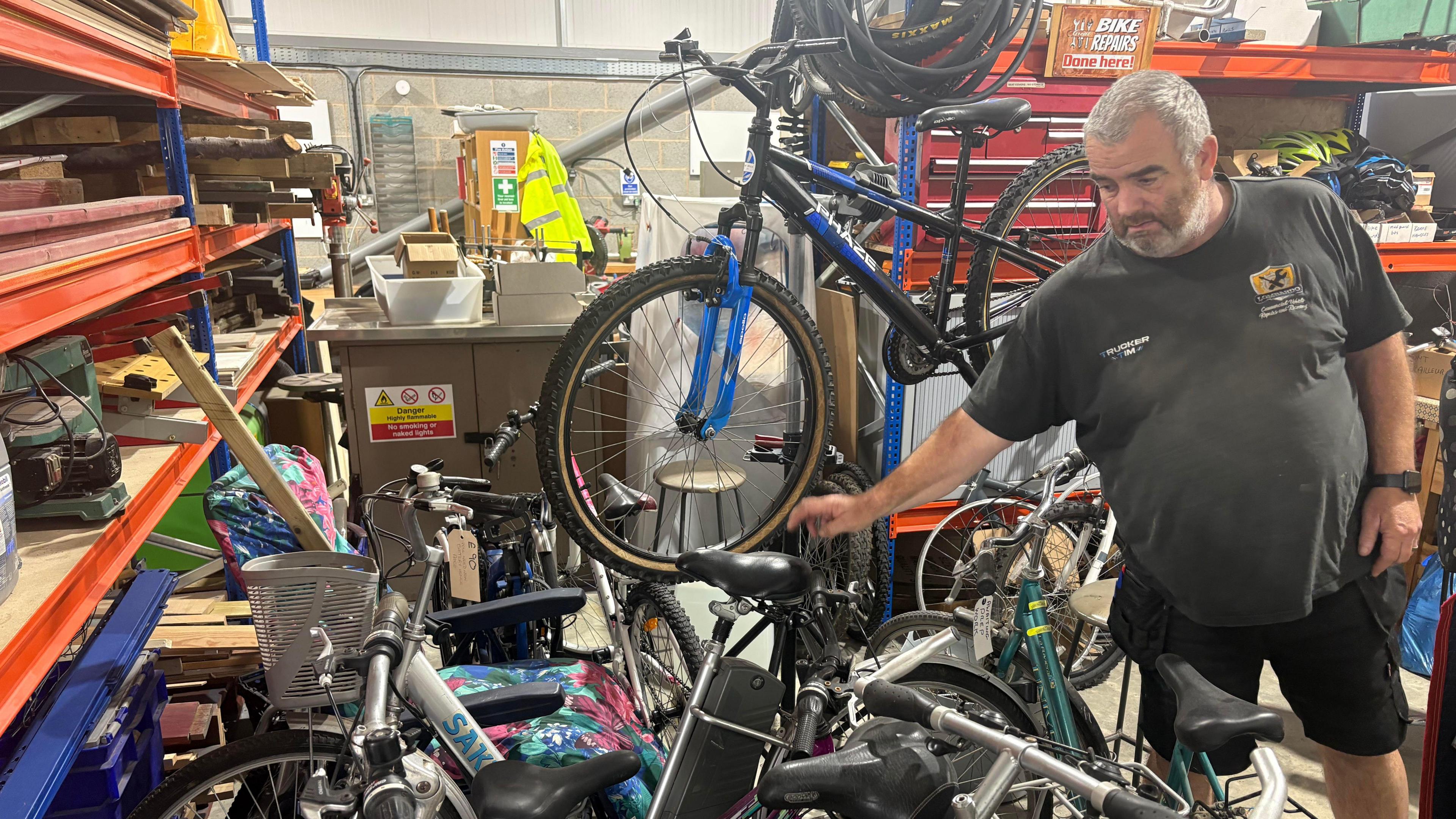 Craig Brown stood among tools, repair equipment and bikes pointing to an electric bike. He is wearing a black t-shirt and black cargo shorts.