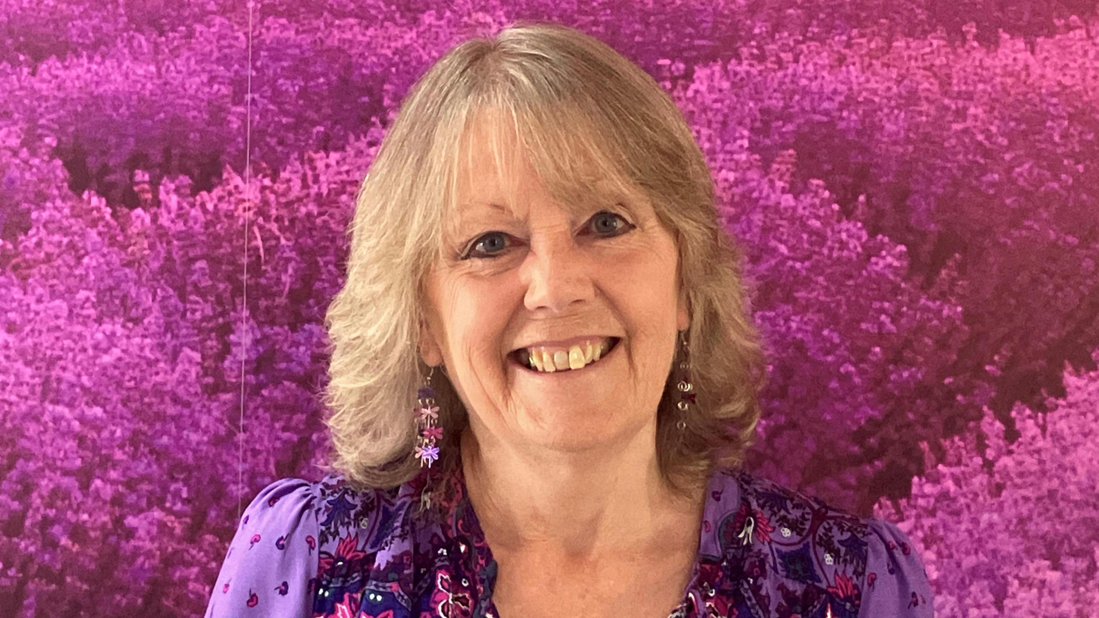 A woman wearing a purple dress and earings, with shoulder length hair, smiling at the camera. She is stood in front of a backdrop of purple flowers.