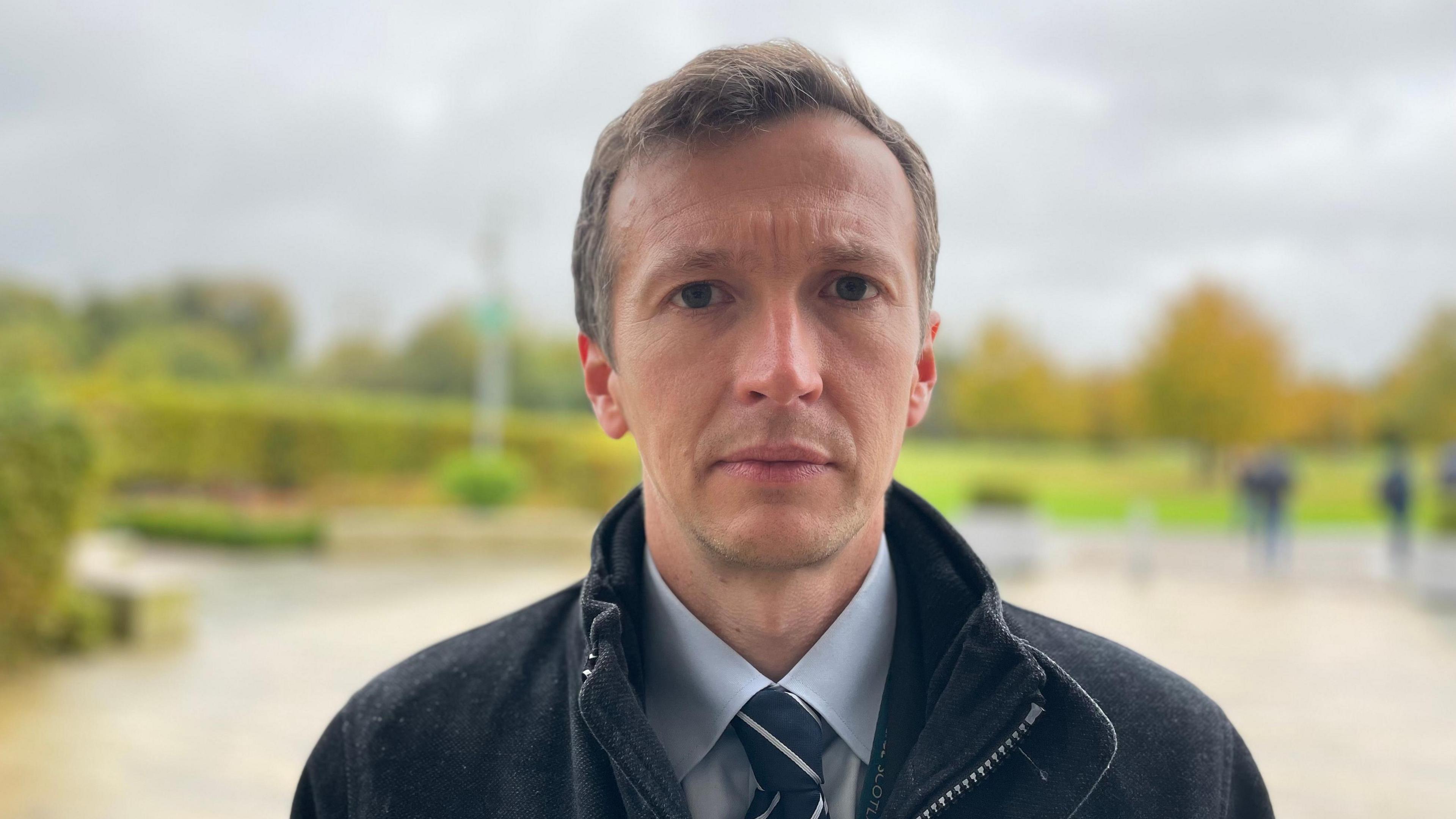 DCI Jonathan Pleasance in front of a blurred background in West Brae, Livingston following a police press conference into the opening of a murder probe after the death of Gary Nicol. DCI Pleasance is looking into the camera wearing a blue shirt with a blue and white striped tie under a dark jacket.