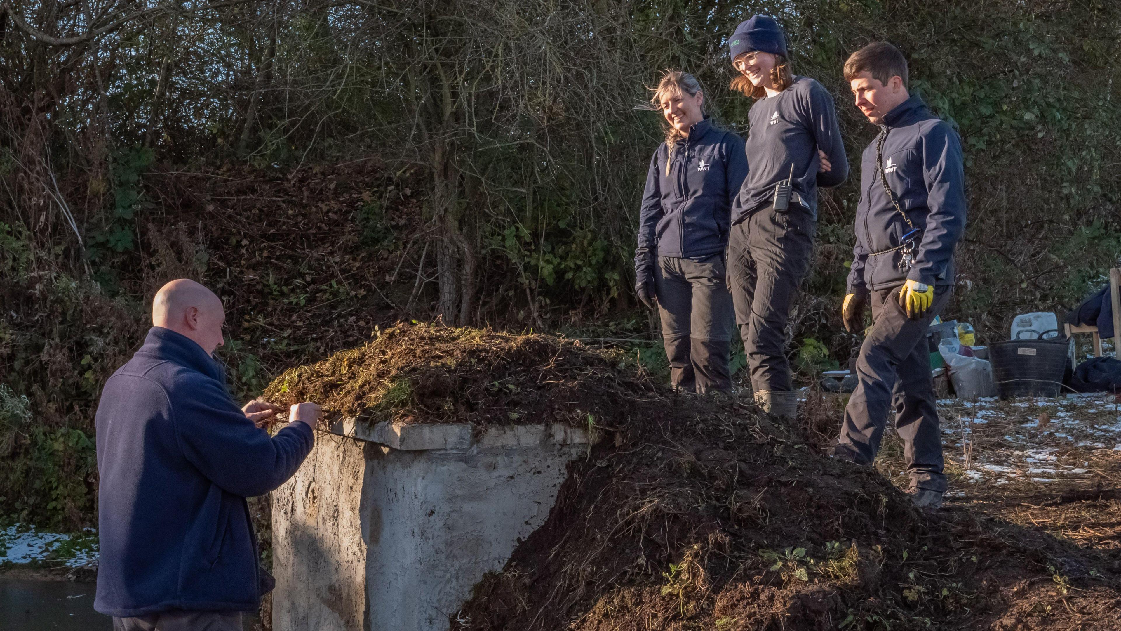 A concrete-looking wall with soil surrounding the structure. A man is touching the soil on top of the wall while three people are standing on top of it looking down.