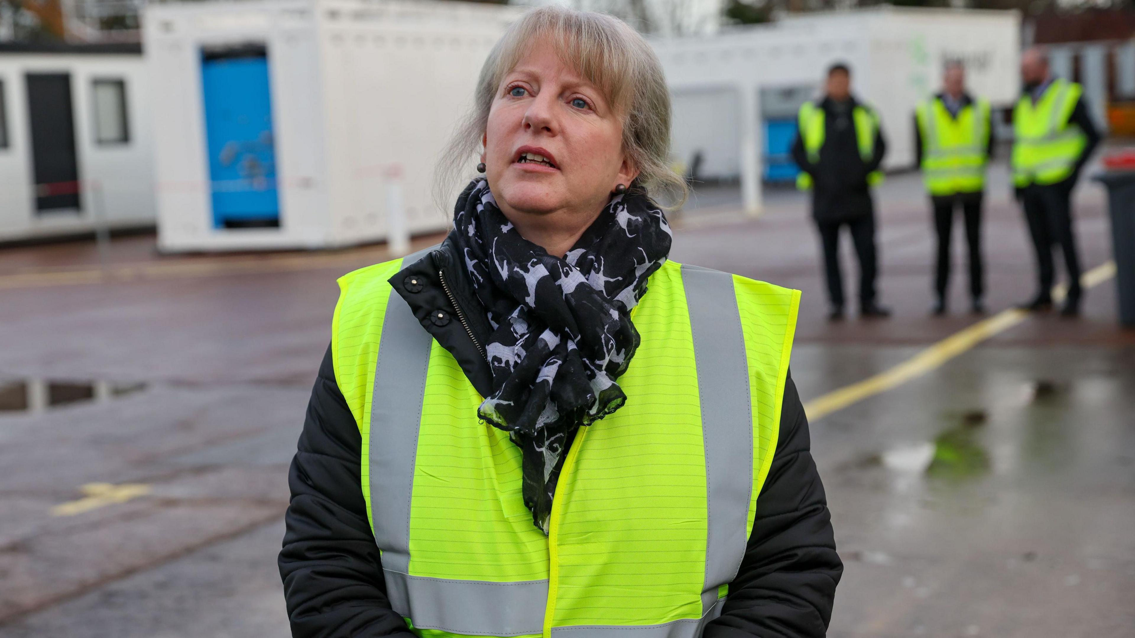 Shona Robison in a green hi-vis tabard and outdoor clothing is standing in an industrial setting. Some men in the same stand behind her.
