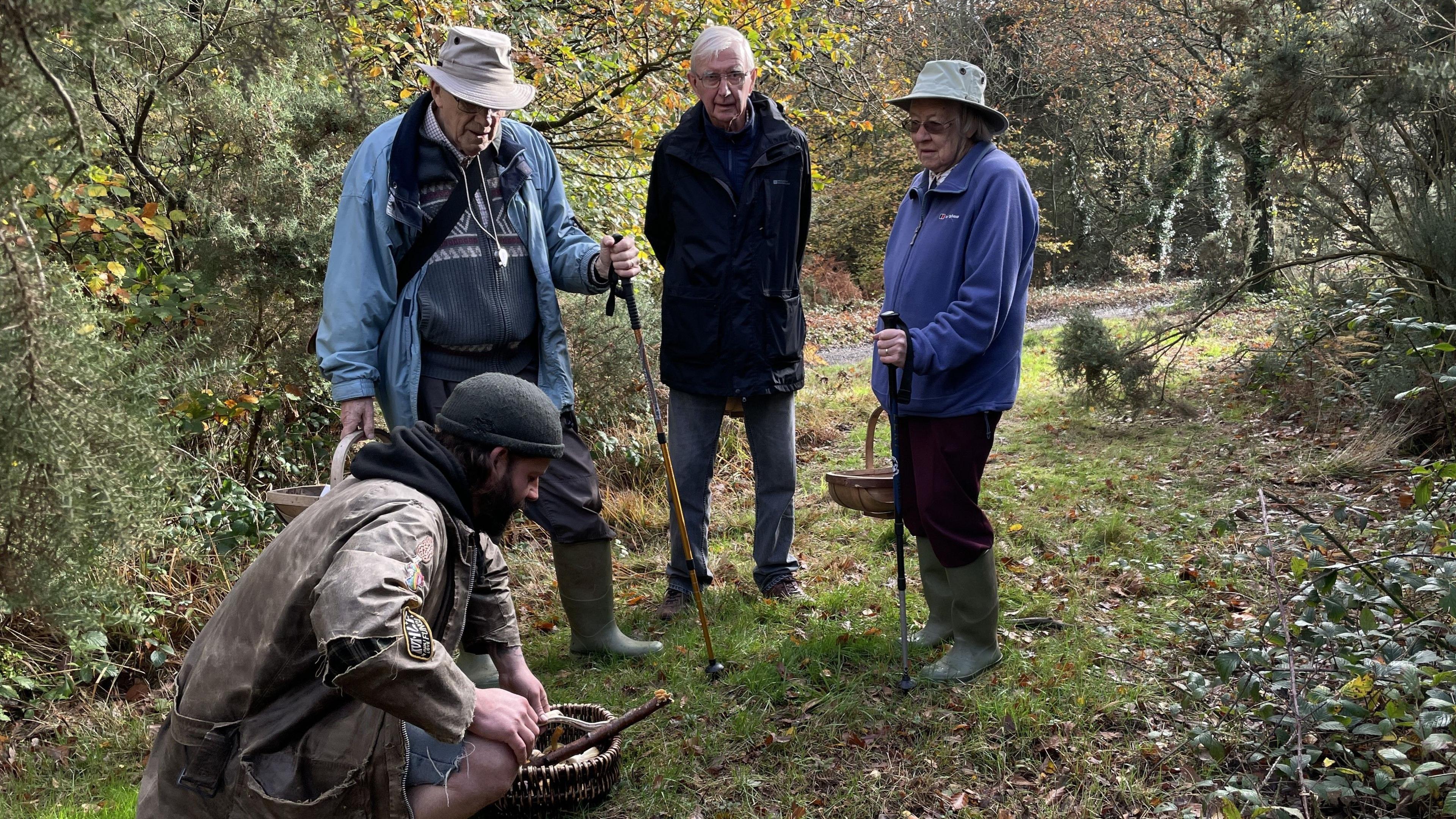 In a clearing in a wood, three people stand around a man giving information about a mushroom he has found. He is crouching down. The others are listening intently. They are all wearing outdoor clothing and carrying baskets. 