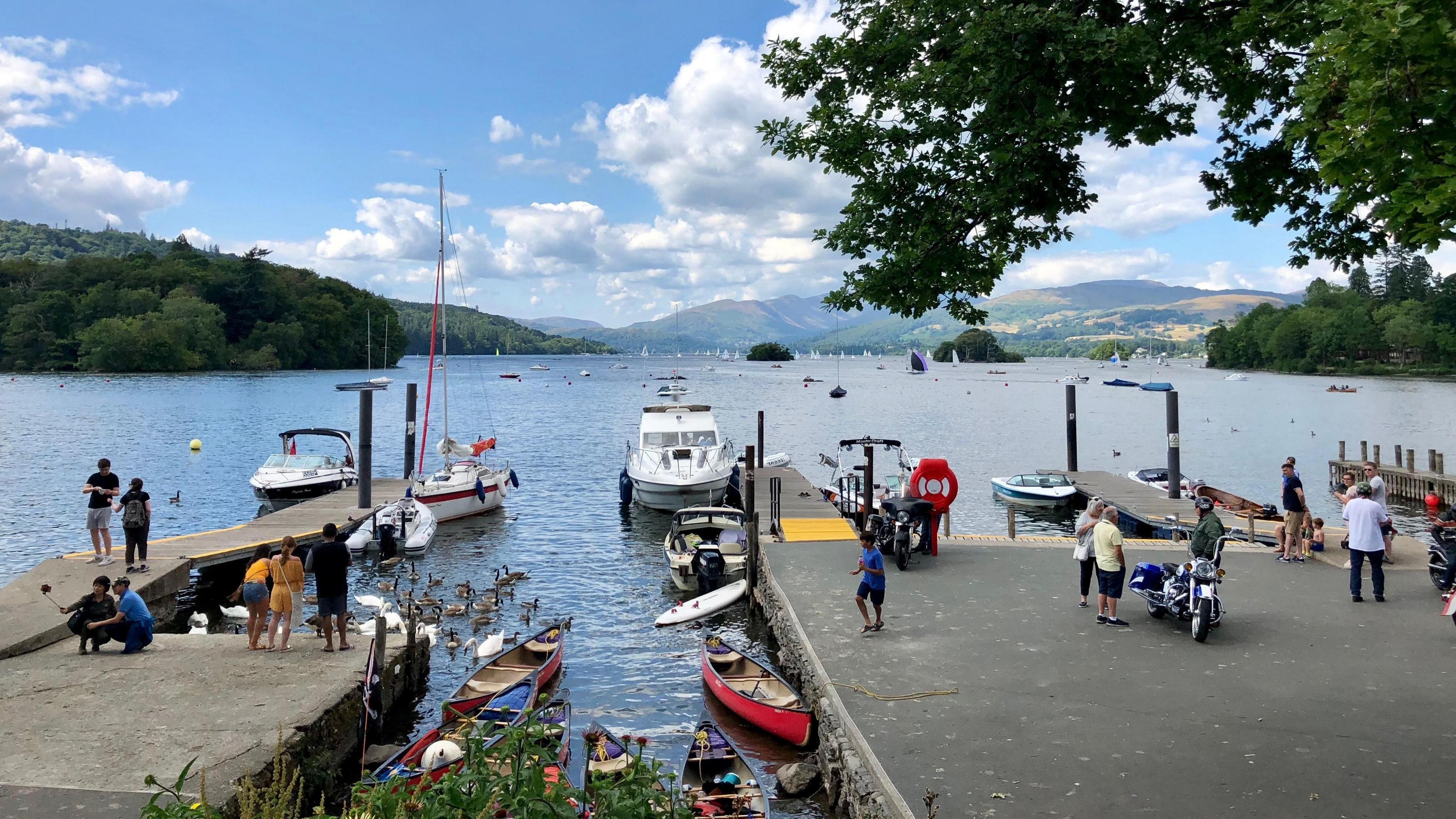 A general view of The Glebe in the Lake District, which shows a lake, people and boats. There are hills in the background.