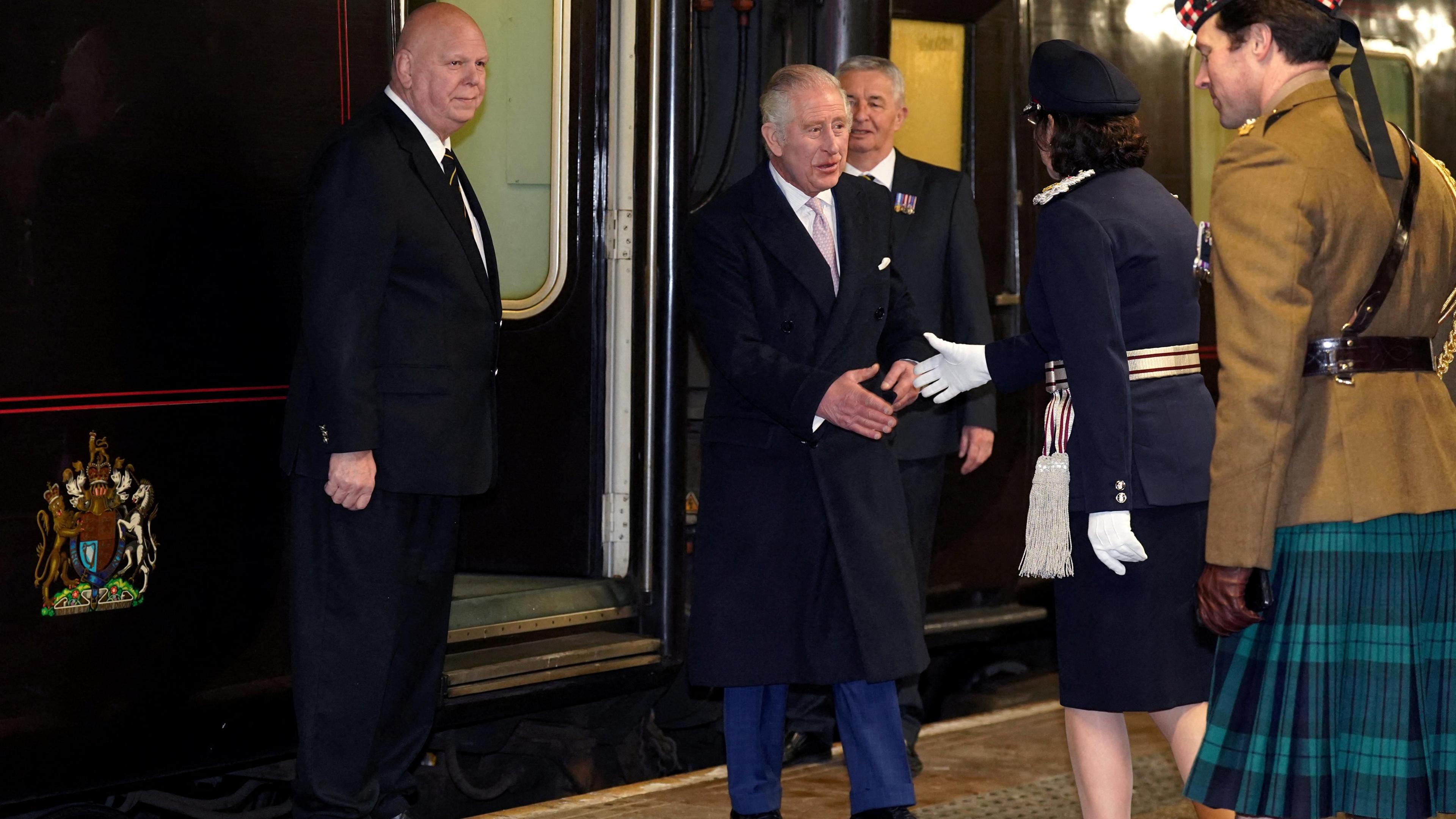 King Charles III is exiting the train and about to shake hands with a uniformed lady on the platform