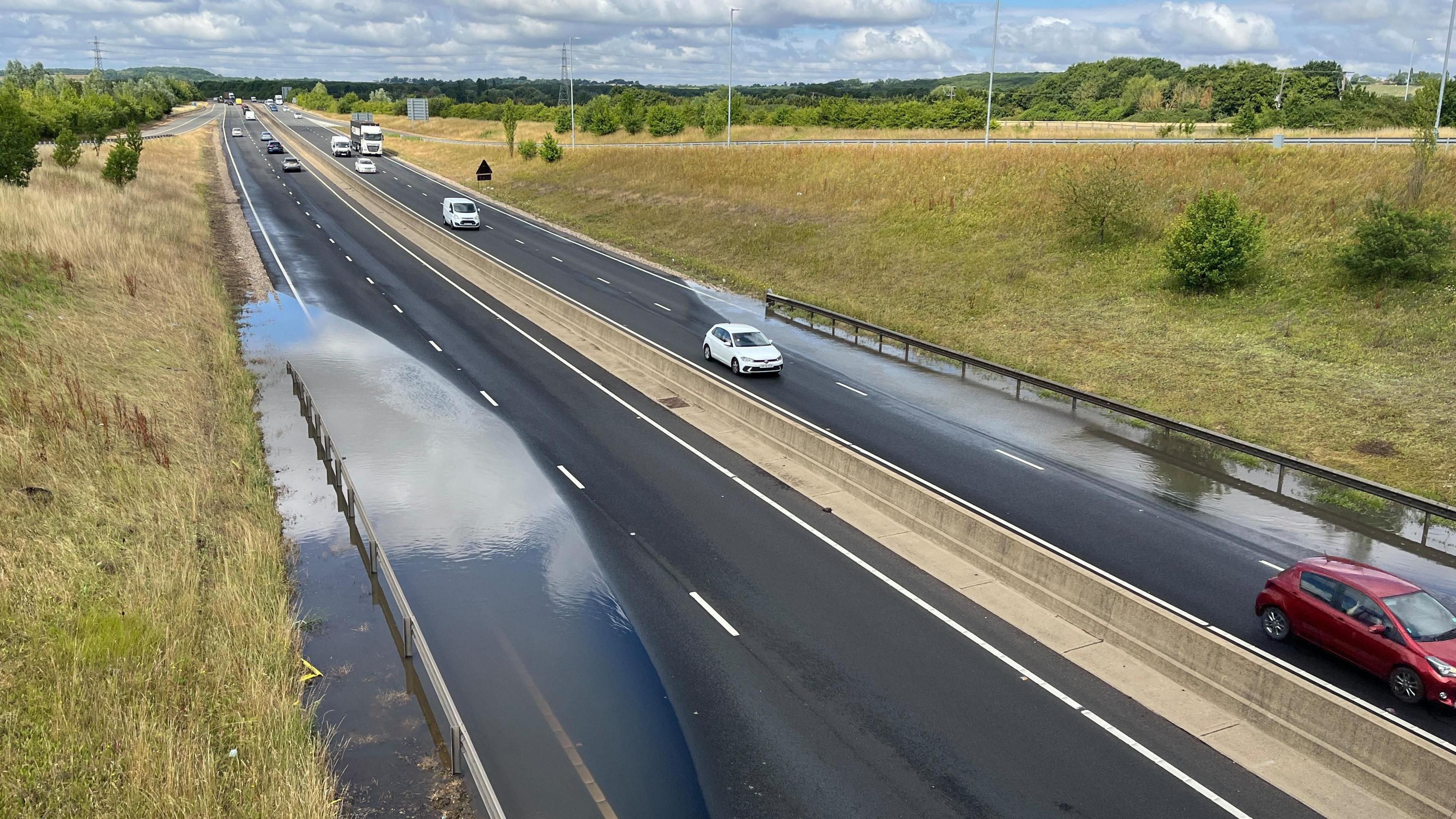 A dual carriageway which is flooded on the nearside lanes on both the east and westbound sections. There is traffic on the road which has trees and grass on either side.