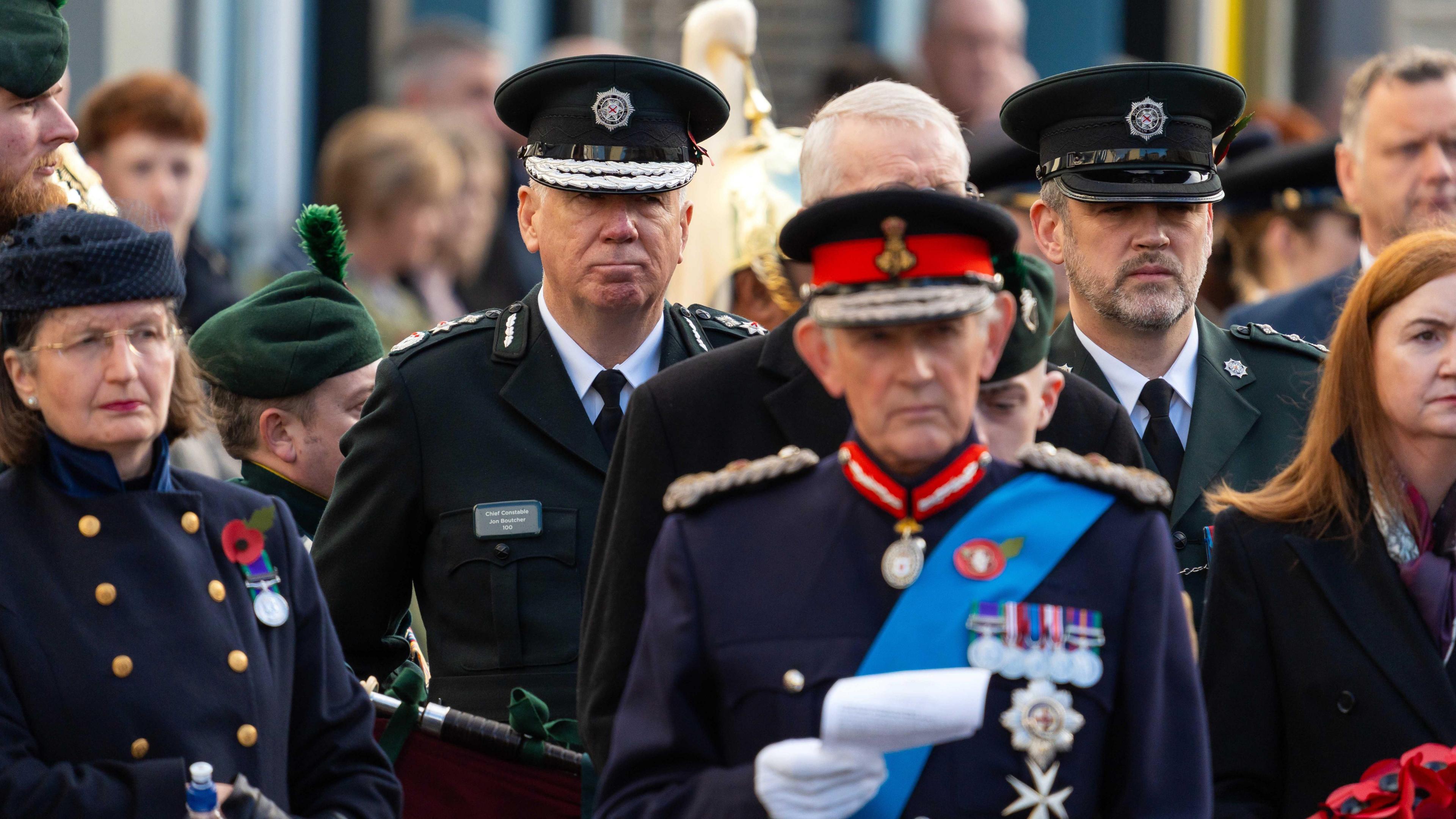 A crowd of people are standing close together. The Chief Constable is wearing his PSNI uniform with lapels on his shoulders. Others are wearing army uniforms. A woman is wearing a navy coat with gold buttons and a poppy.