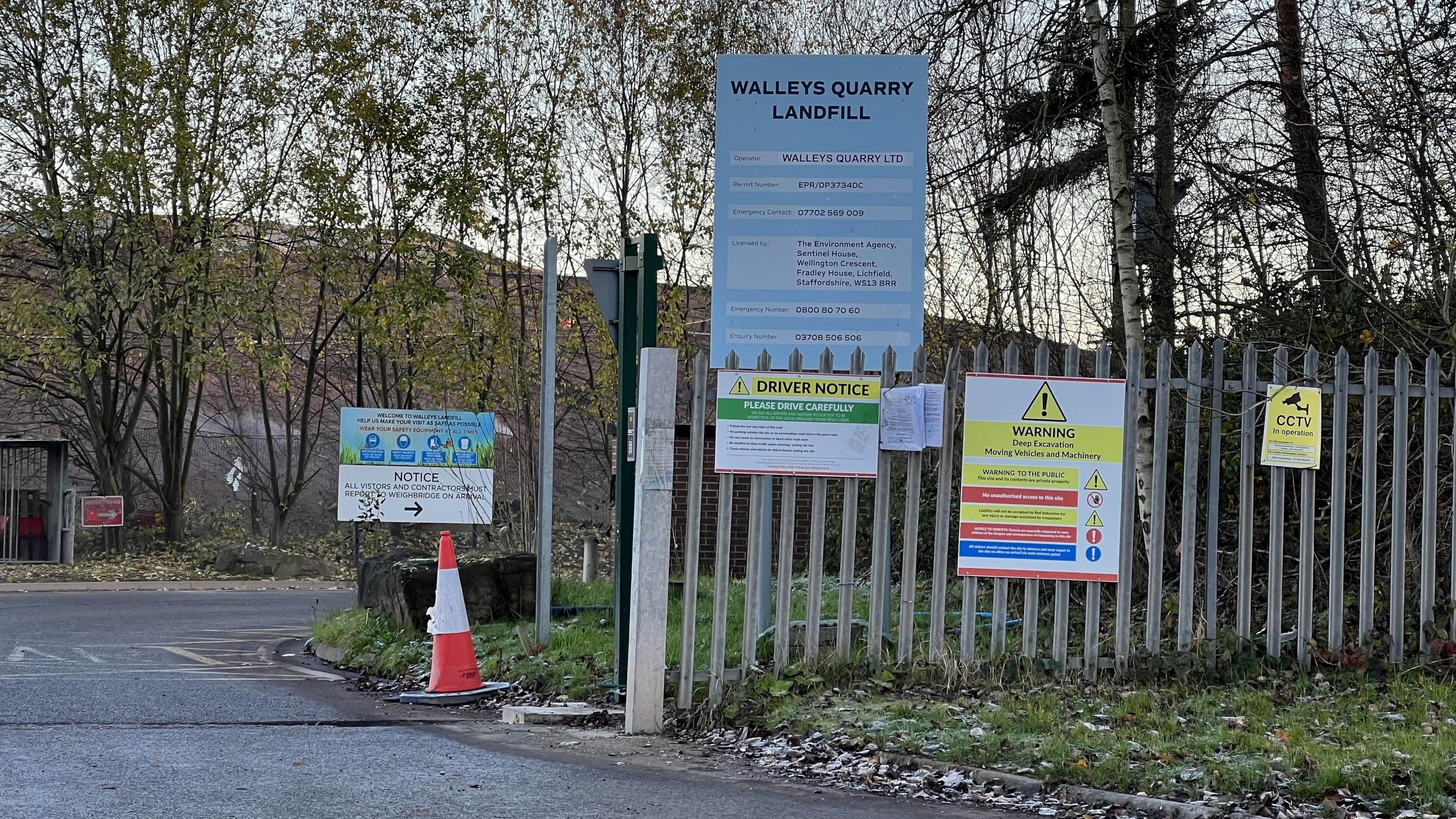 The entrance to Walleys Quarry  -  a metal fence on the side of a road has a blue sign attached to it that says "Walleys Quarry Landfill" along with other warning signs. A landfill is barely visible in the background behind a row of trees.