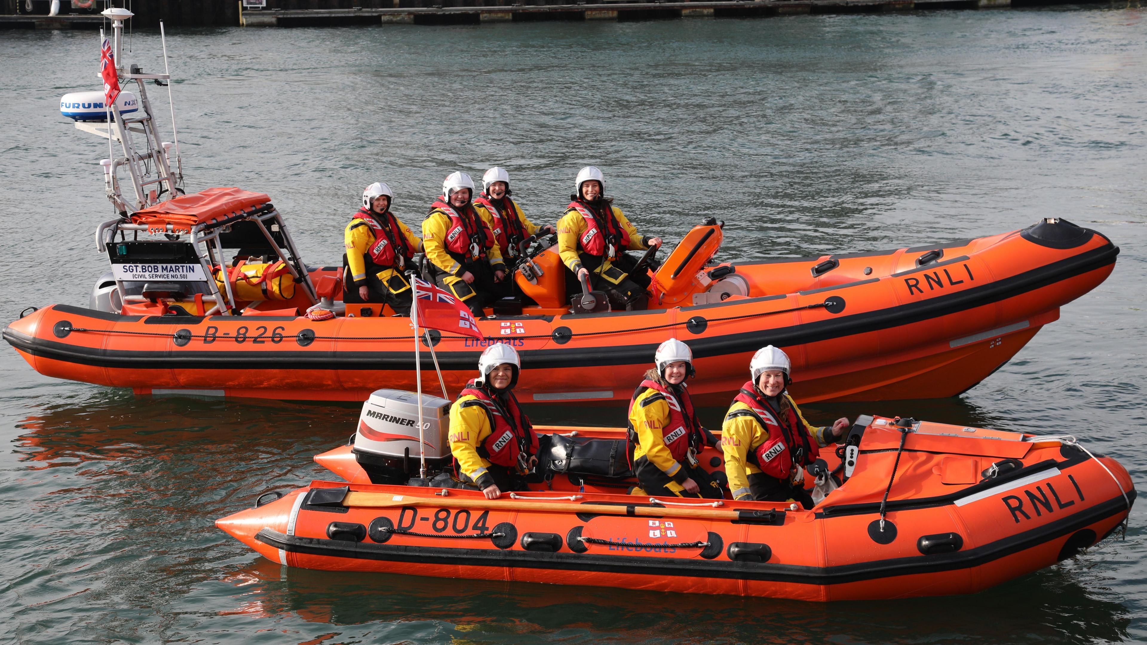 Two RNLI lifeboats sit side-by-side in the water. There are four crew member in the large boat and three in the other. They are wearing white helmets and are looking at the camera.