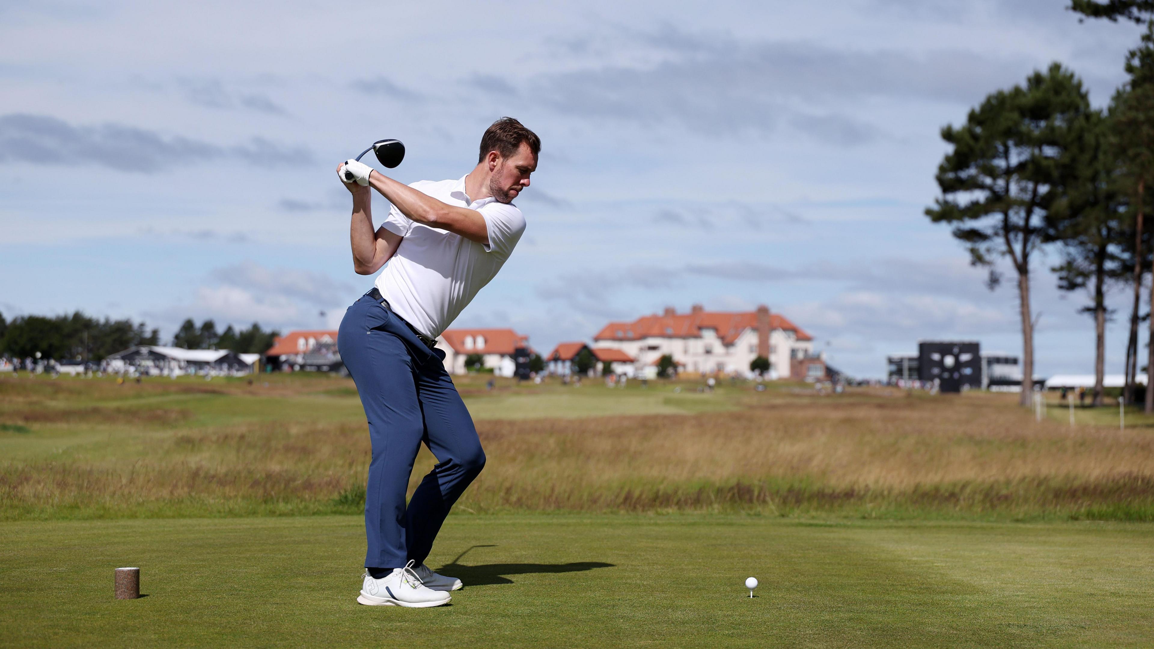 Steven Whittaker, pictured at the top of his back swing, tees off on the second hole during the pro-am. Houses can be seen in the blurred background ahead. He is wearing a white polo shirt, blue trousers and white golf shoes.