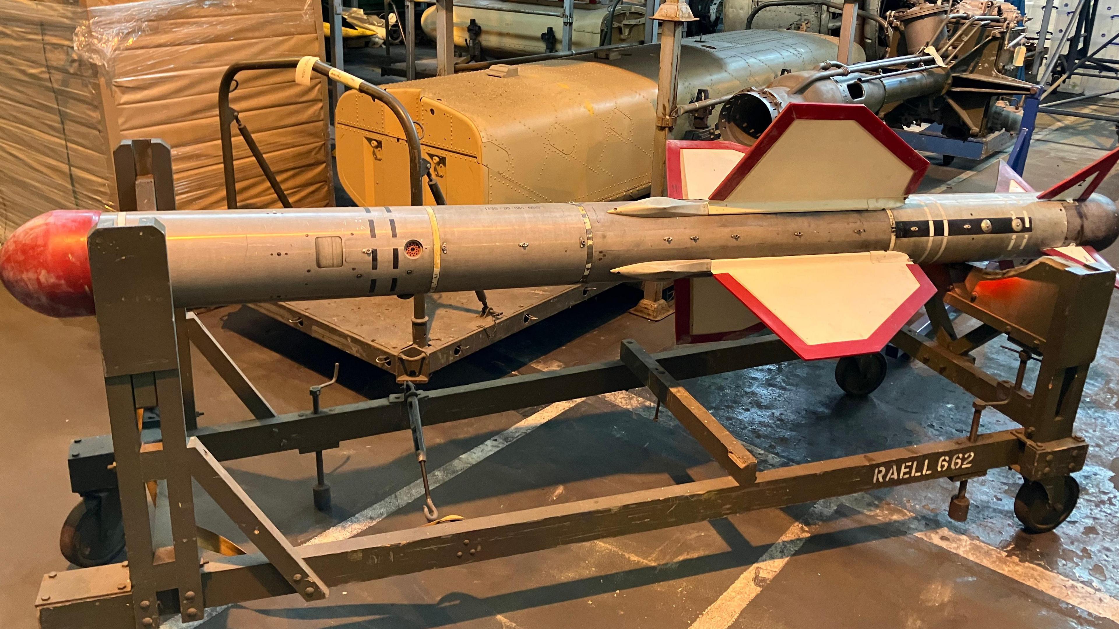 A silver and red deactivated RAF missile on a green, metallic stand with wheels. Behind it are shelves with boxes placed on them.