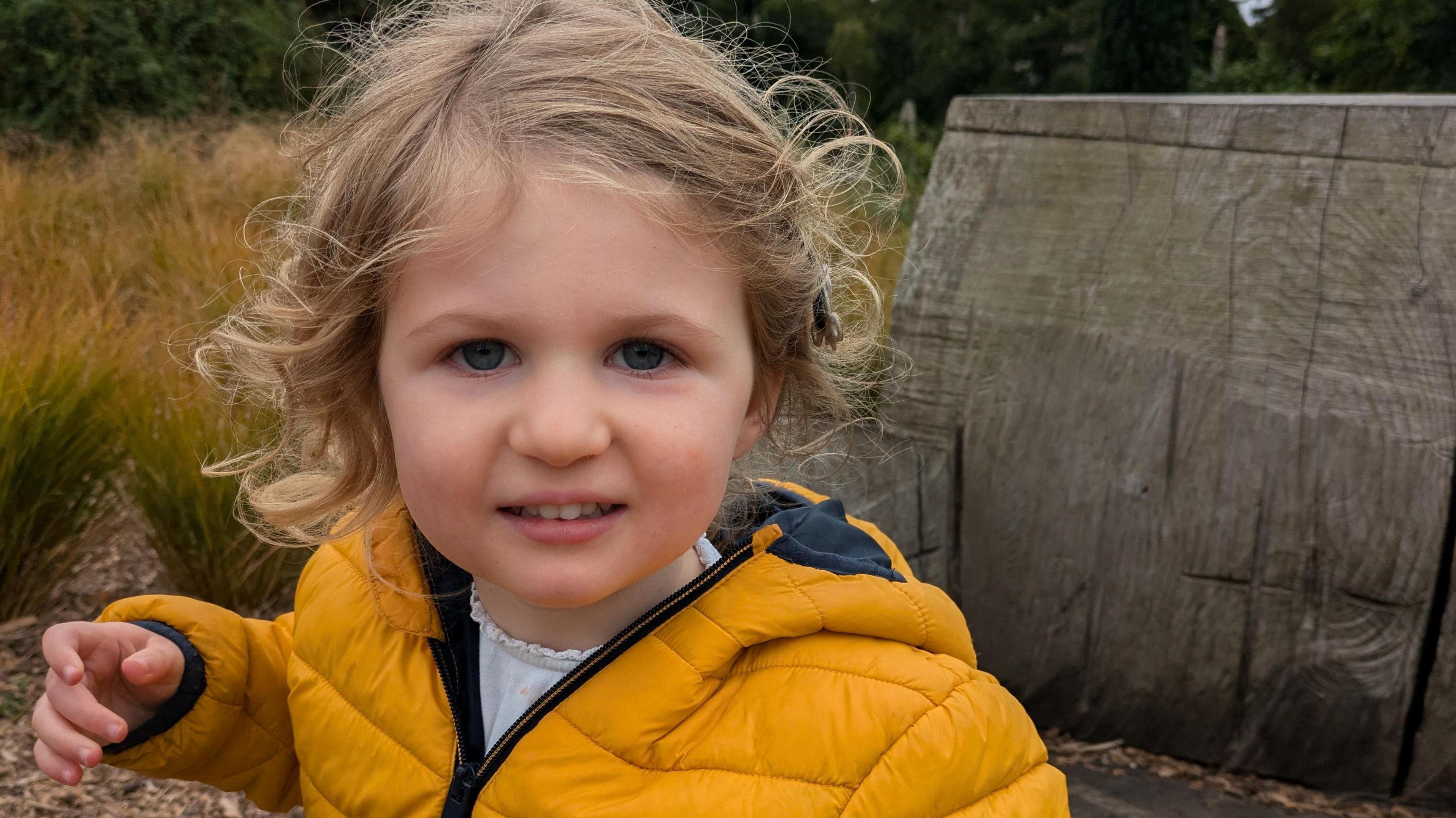 Joy is wearing a mustard colour winter jacket. She is outside in a field or woodland scene. There is a seat or bench behind her that has been carved out of wood.