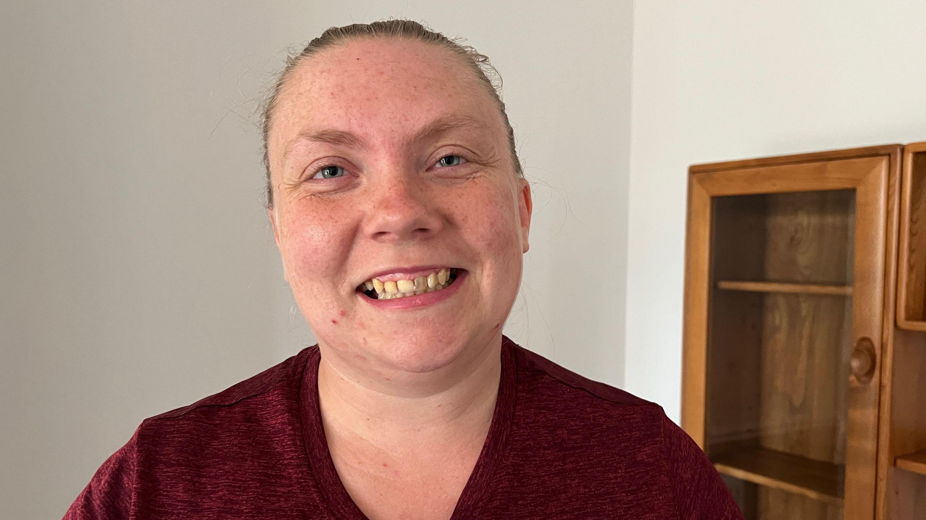 A woman who is smiling at the camera has her fair hair tied back and is wearing a burgundy T-shirt as she stands in front of a wall cabinet.