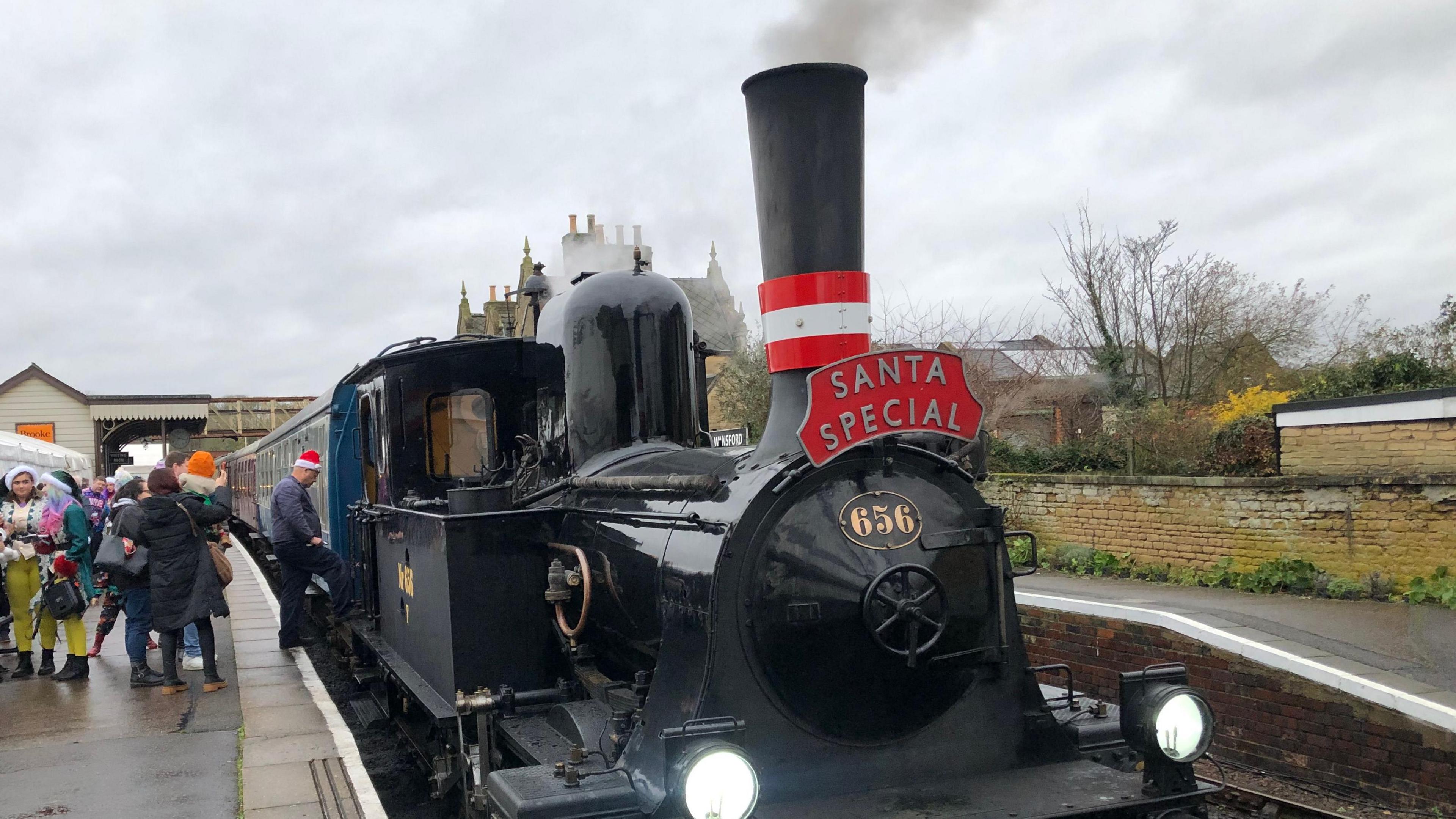 A black steam engine with a plate containing gold numbering, saying "656". Smoke is coming from the engine's chimney and a red plaque says "Santa Express" on it. People can be seen boarding the train further along the platform