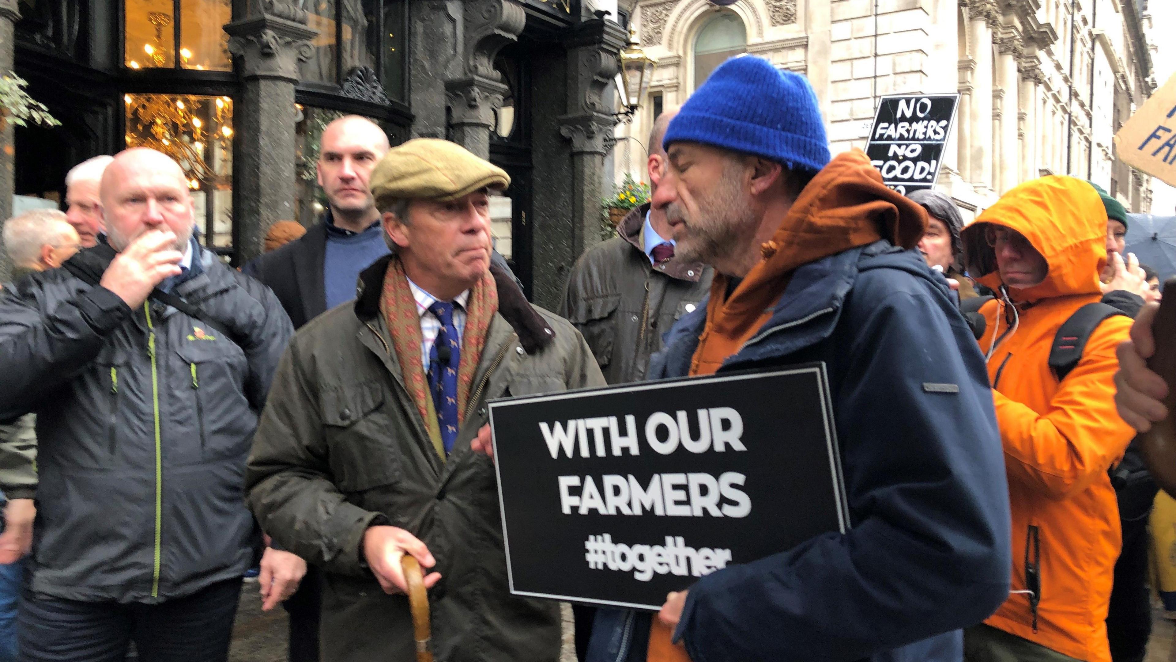 A protestor holds a banner saying 'With our farmers #Together' while talking to Nigel Farage. A number of people are around them on a street in London. Another placard seen in the background says 'No farmers! No food!'. A man appears to be filming Nigel Farage and the protestor next to him. 