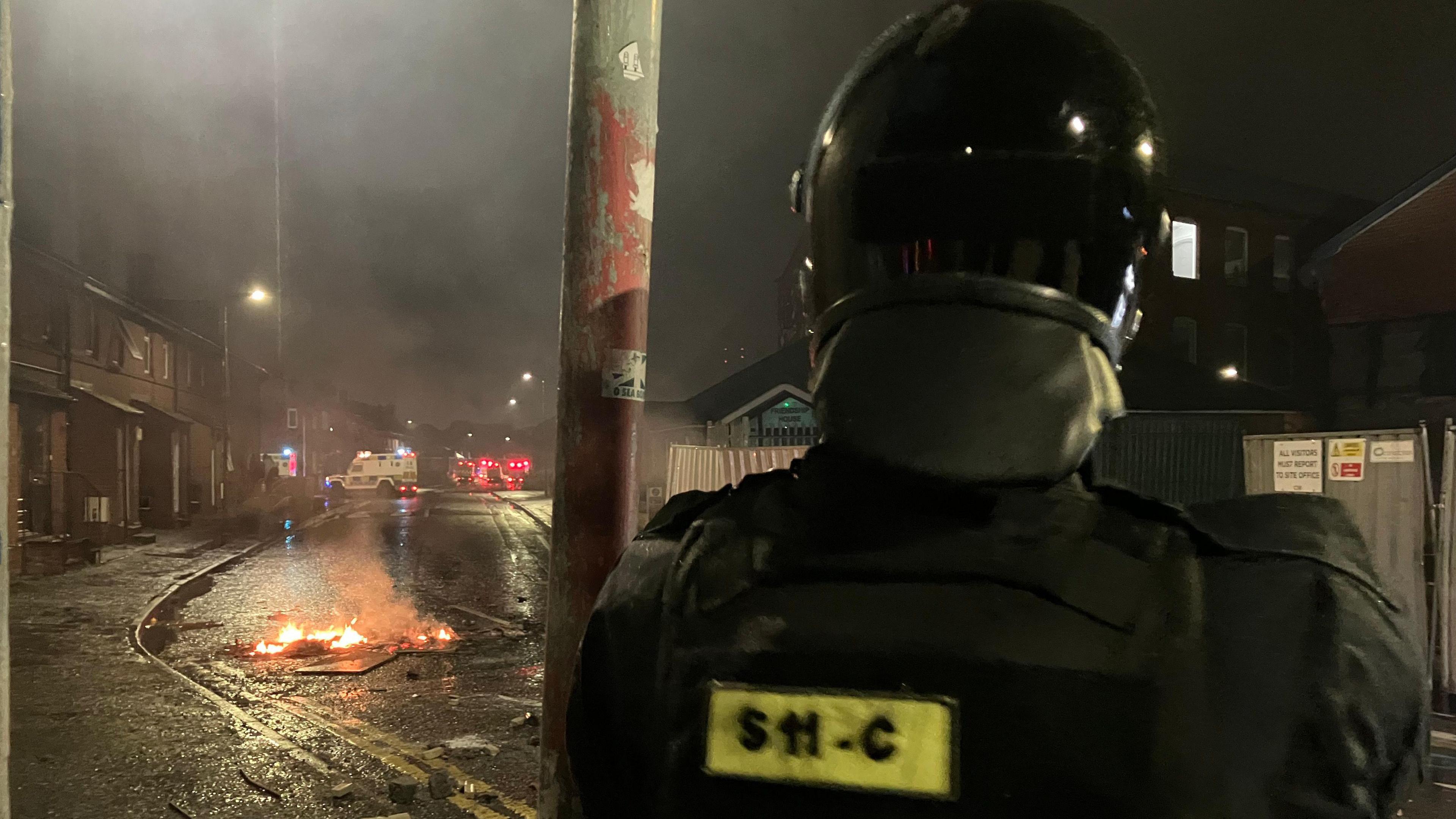 A police officer in riot gear looks on as something burns in the middle of the road. In the Background a police landrover