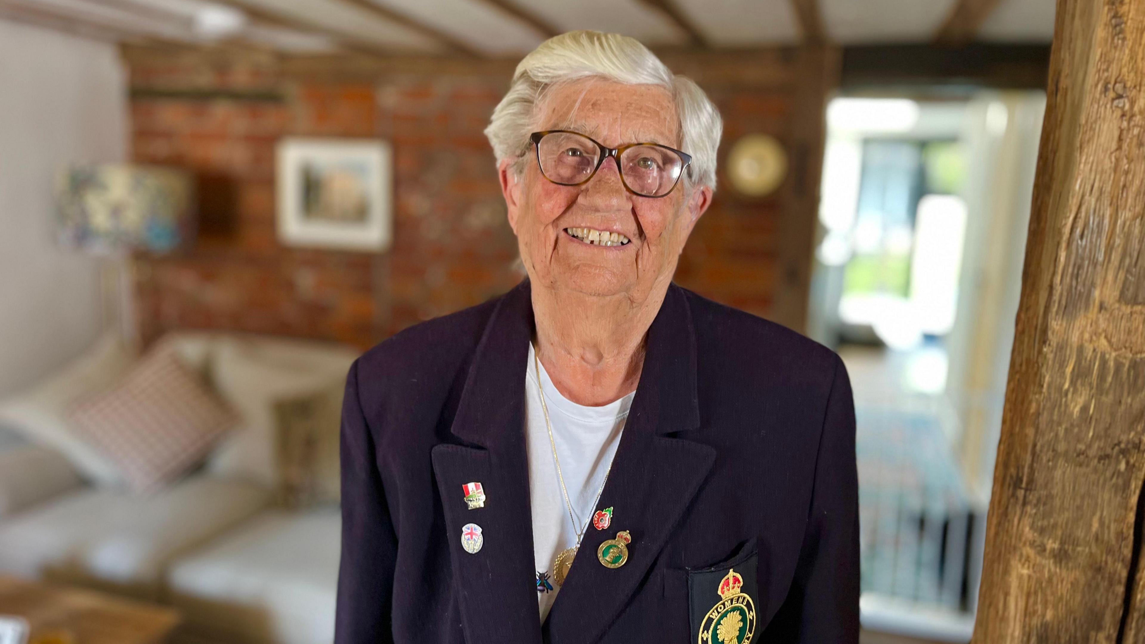 Lady with silver hair and glasses smiling into the lens. She is wearing a white t-shirt and a navy blazer with badges either side, and a Land Army logo on the breast pocket.
