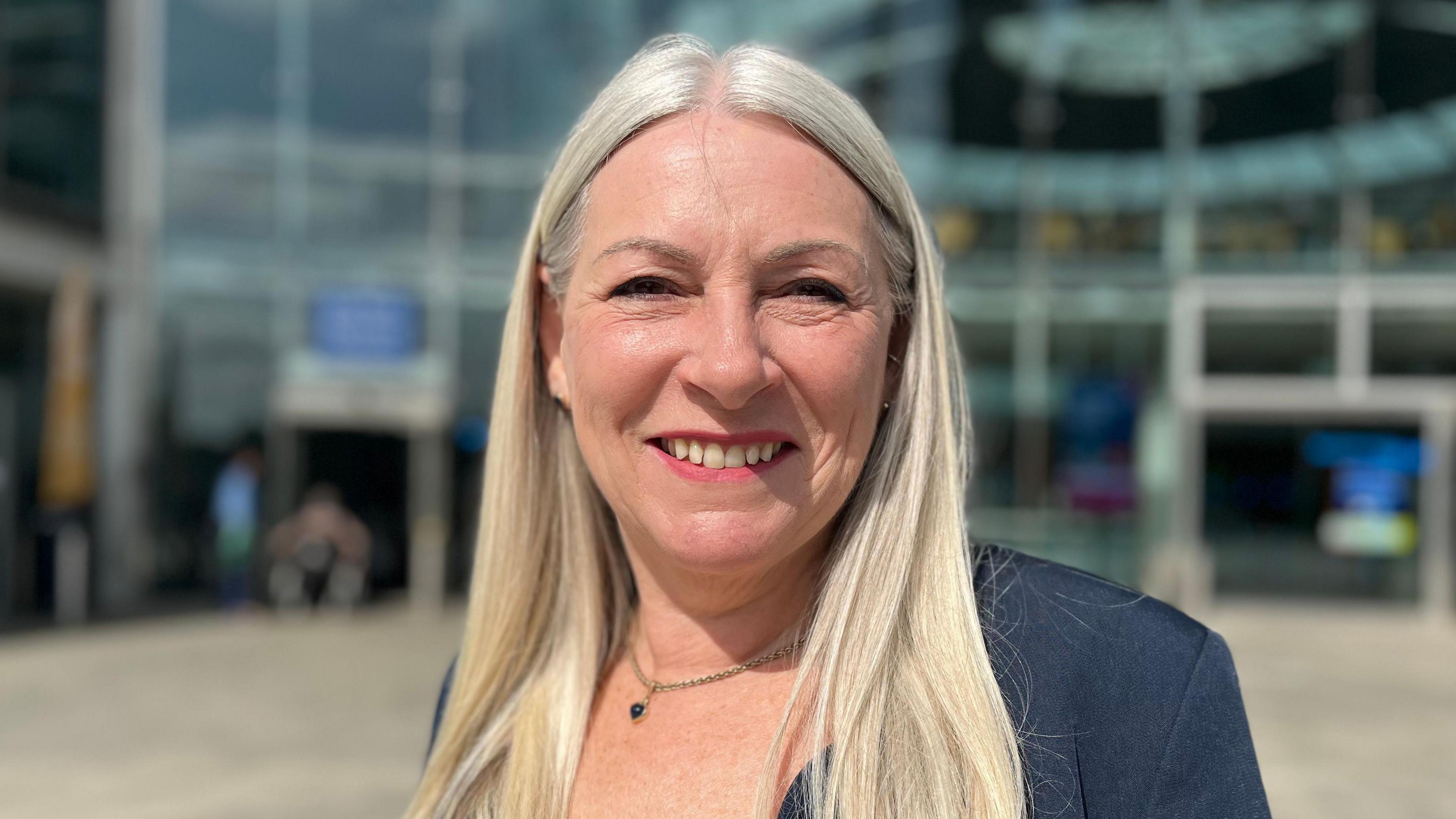 Kay Mason Billig smiling and wearing a dark blue suit jacket. It is clearly sunny weather. She is standing in front of a glass-fronted building. 