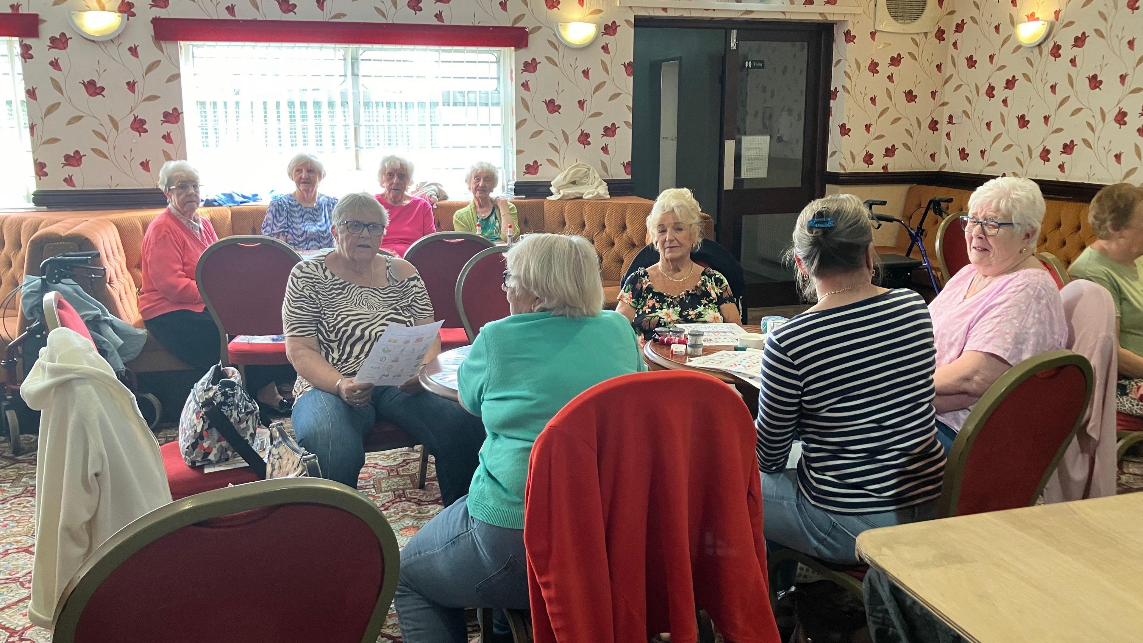 A group of older women take part in activities at their social club. They are sitting around tables in groups, looking at pieces of paper.