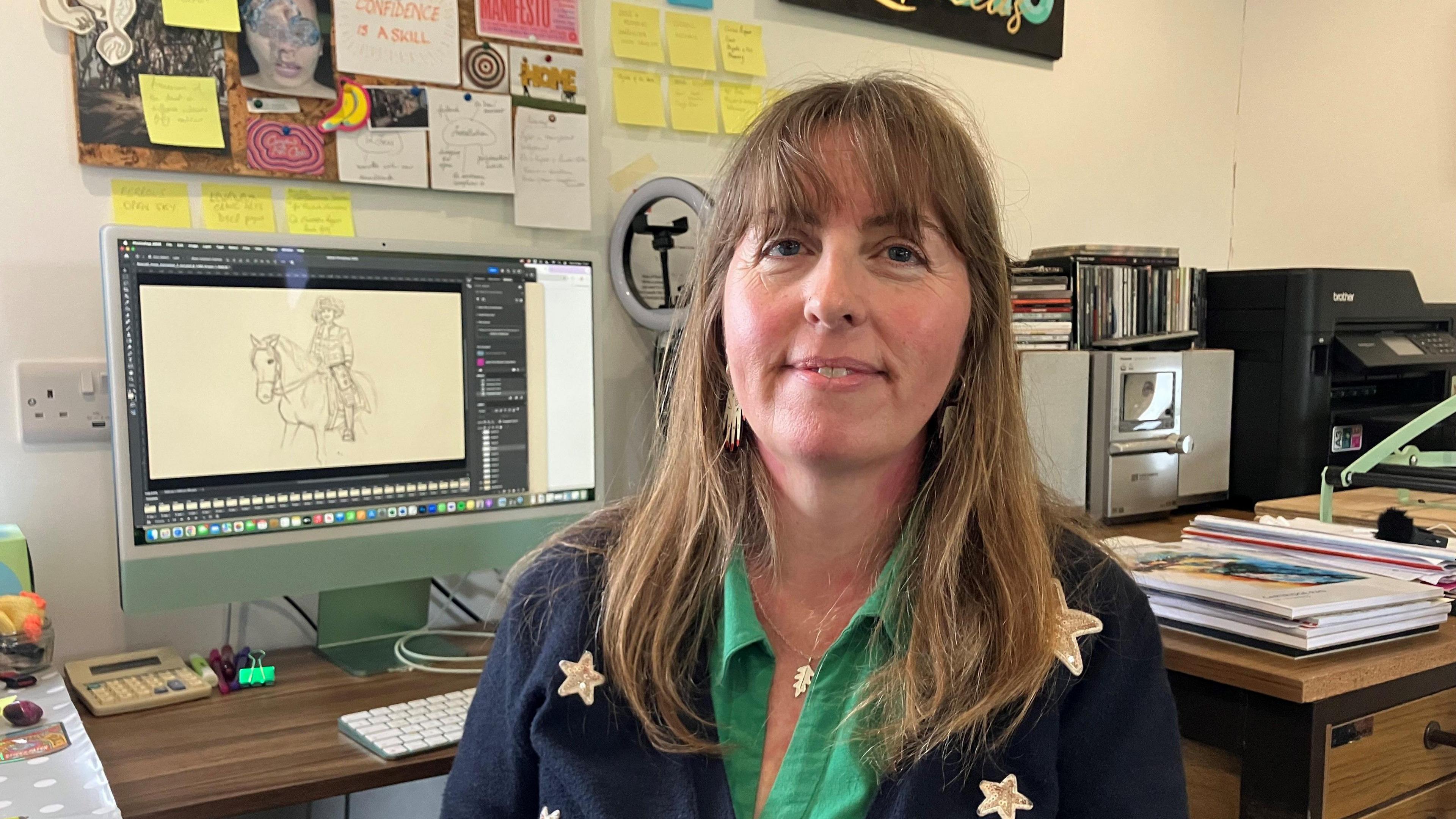 Rebecca Farkas sits in front of her work computer, with a hand-drawn image of a circus horserider visible on the screen. She is smiling and wearing a green shirt, and dark blue jumper with stars on.